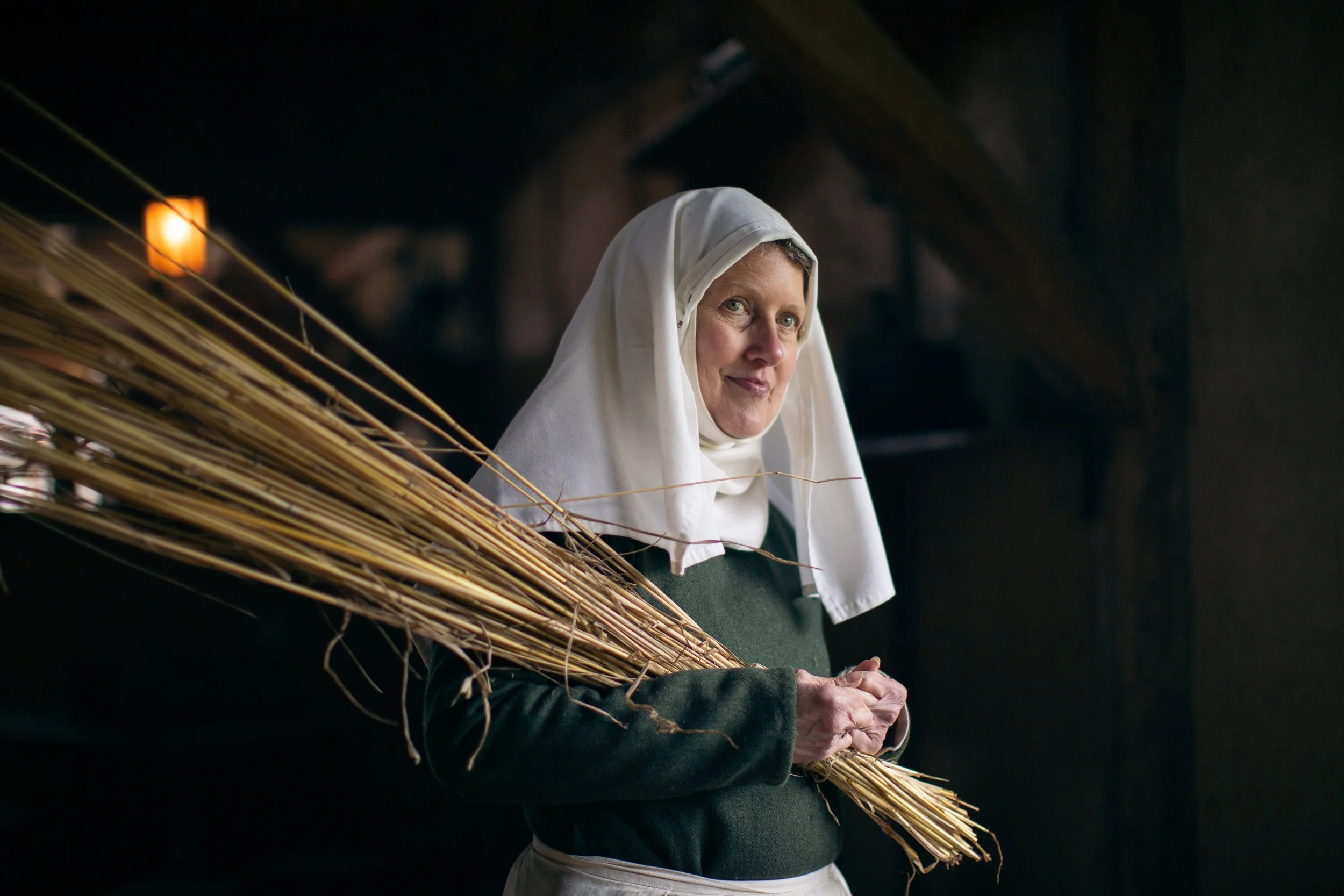 A woman with light skin and gray hair, wearing a white headscarf and black sweater, holding a large bundle of dried reeds or straw in her hands, standing indoors with a dark background illuminated by a small orange light.