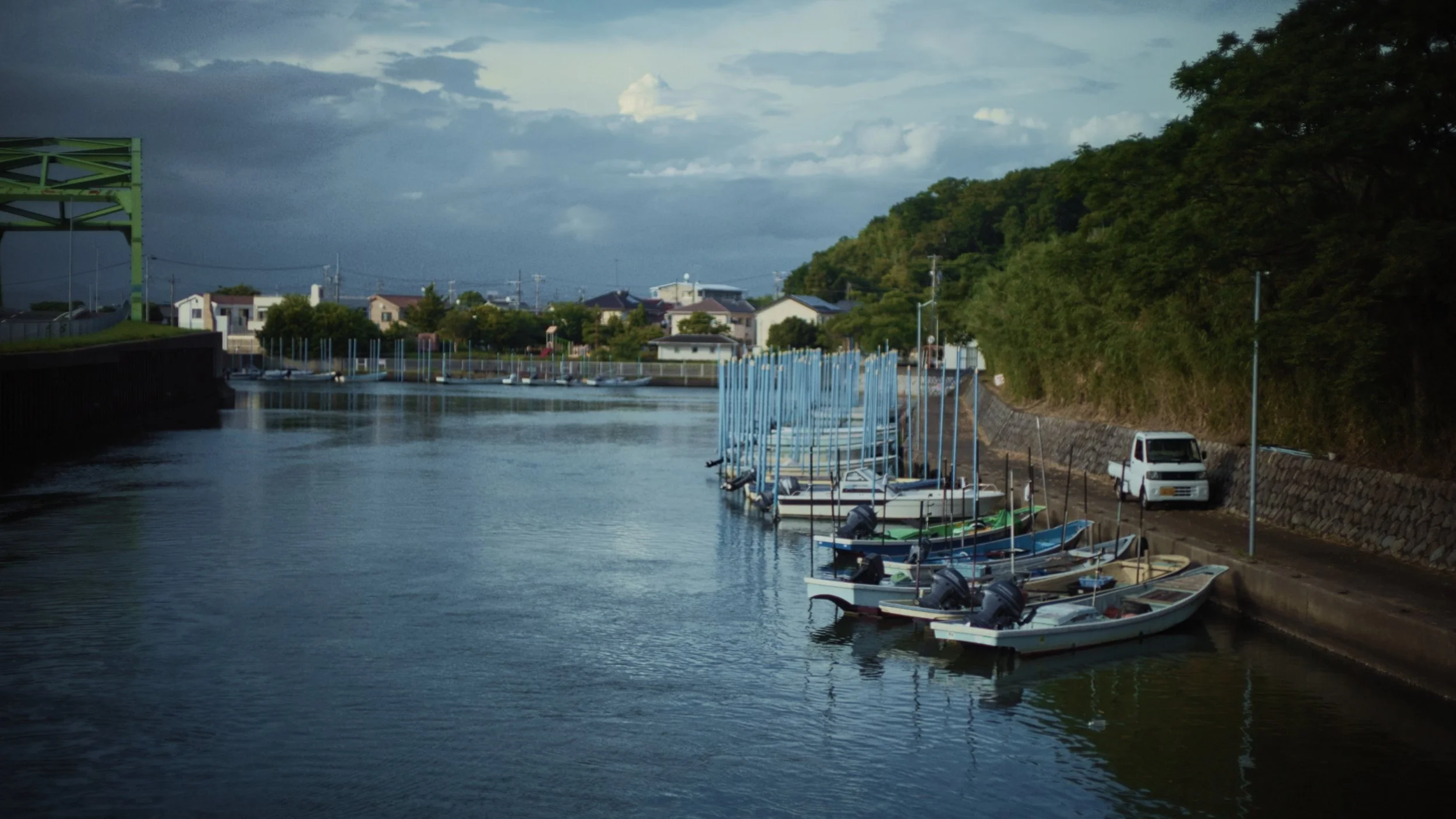 A small marina with boats docked along a narrow waterway, houses in the background, with a hill covered in trees on the right and a cloudy sky overhead.