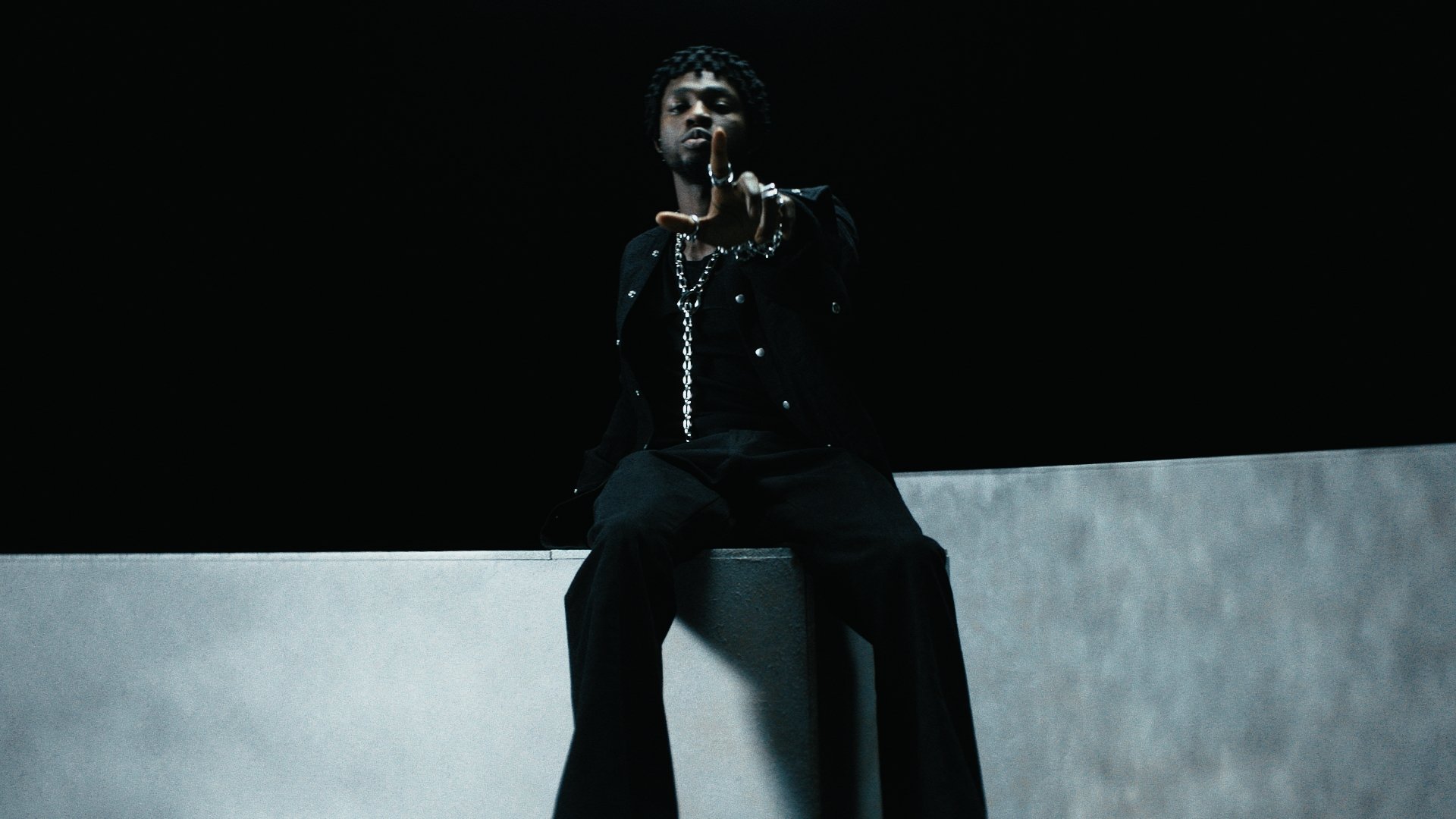 A person with curly hair and dark clothing sitting on a ledge against a black background, with a serious expression and hand extended towards the camera.
