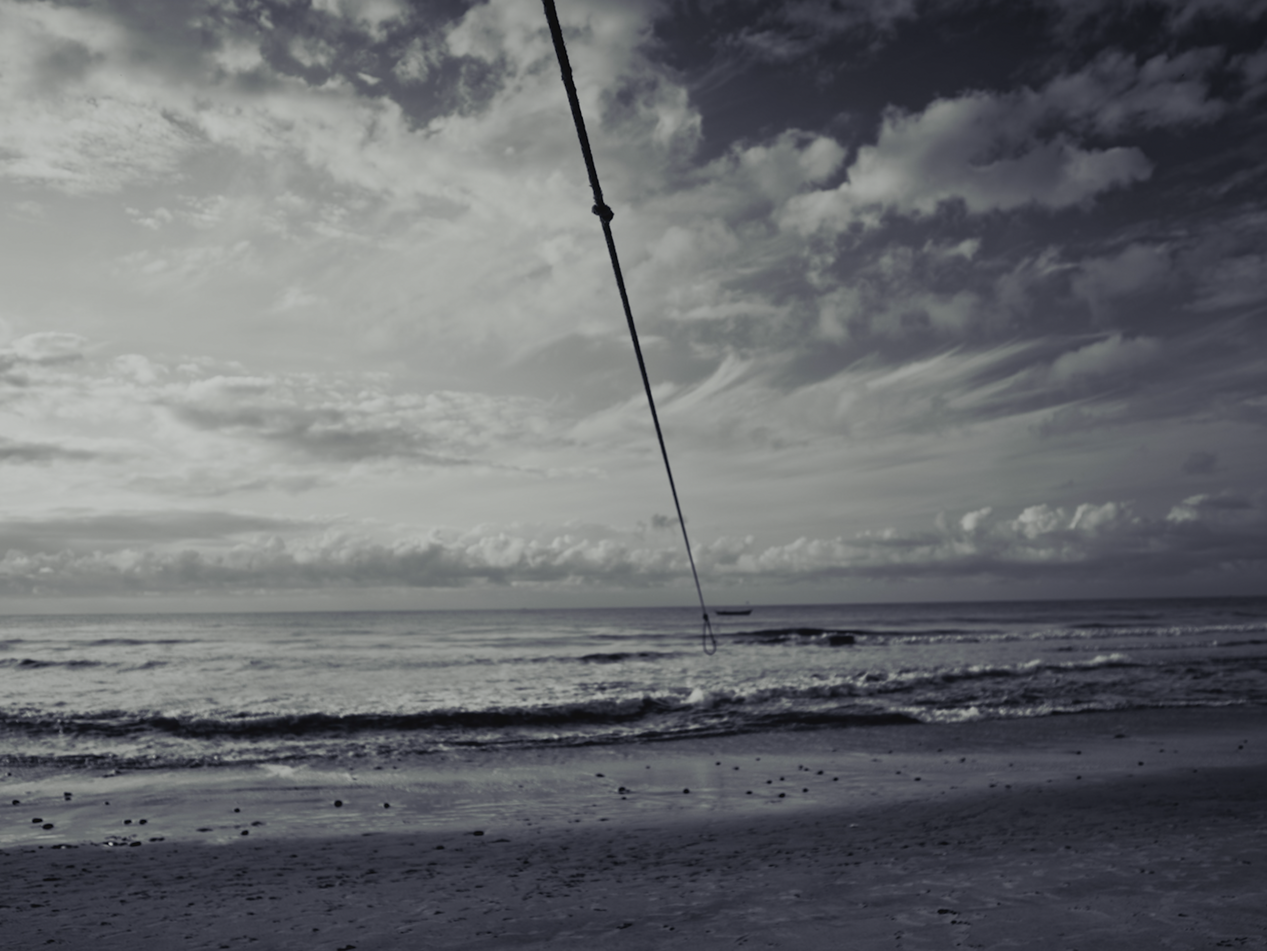 Cloudy sky over the ocean with waves, a shoreline, and a surfboard attached to a rope hanging from above.