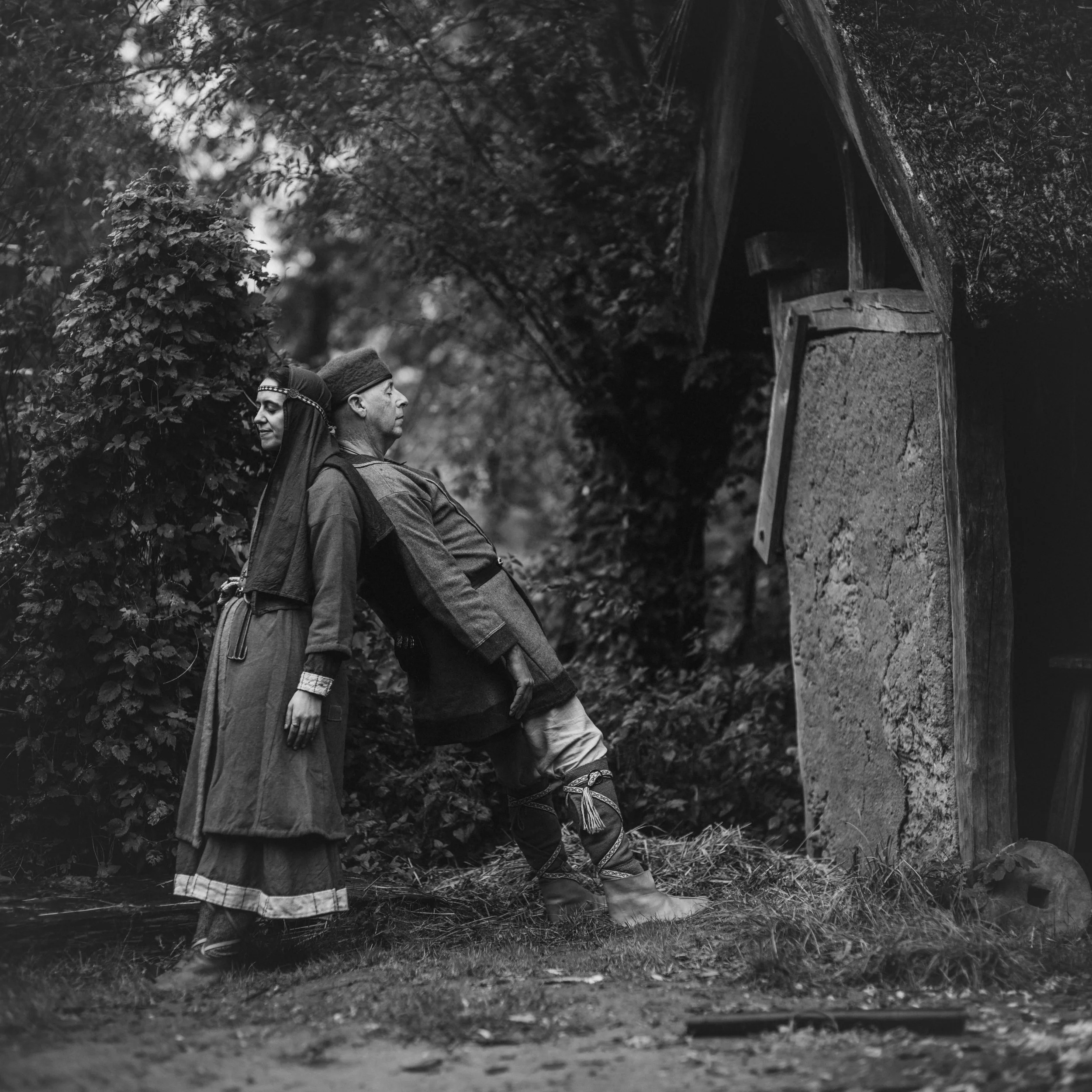 Black and white photo of three Indigenous women dressed in traditional clothing standing outdoors near a dirt path and wooden structure, surrounded by foliage.