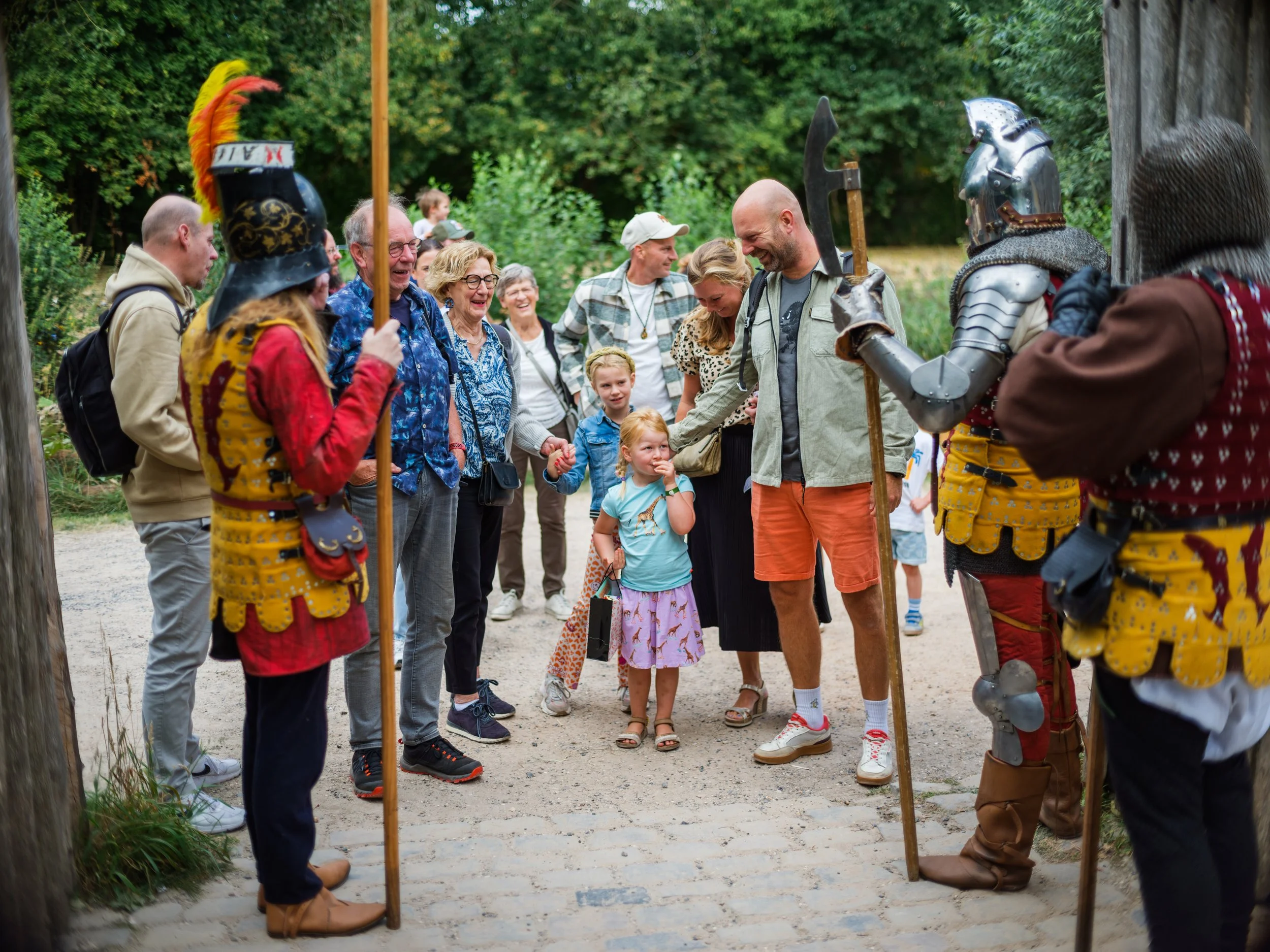 Group of people, including children and adults, with some dressed as knights in armor, gathered outdoors in a wooded area, smiling and engaging in conversation.
