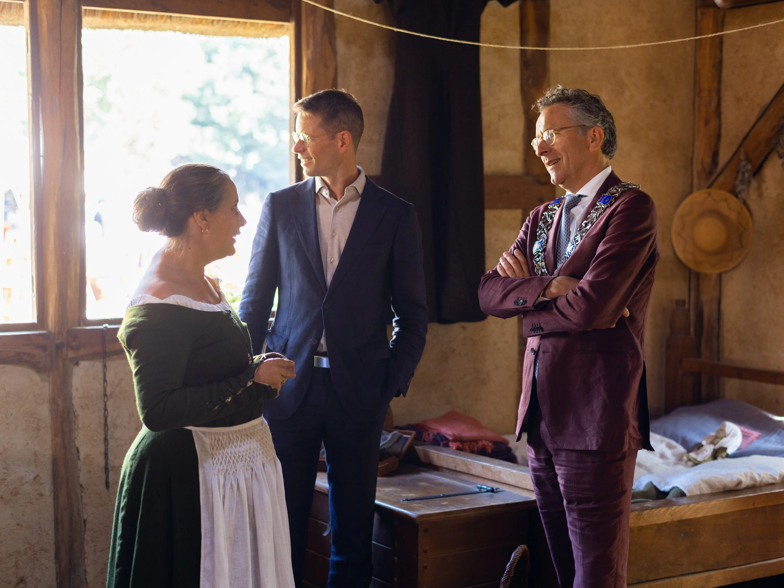 A woman in a white and green dress talking to two men, one in a suit and the other in a burgundy blazer, inside a rustic wooden room with natural light.