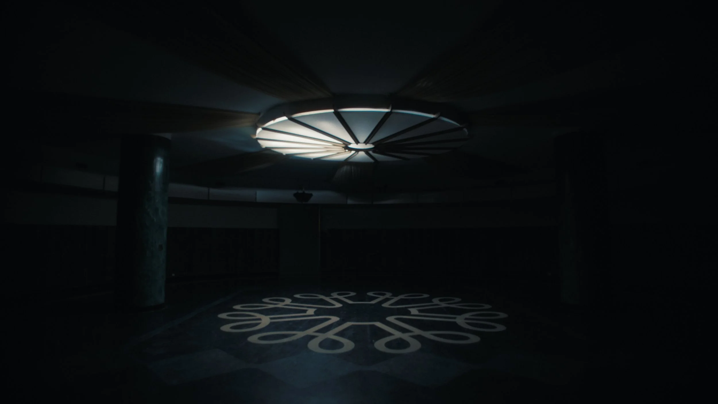 A dark interior space with a illuminated circular structure on the ceiling, featuring radial lines. There is a partially visible pillar and some text on the floor that appears to display a phone number.
