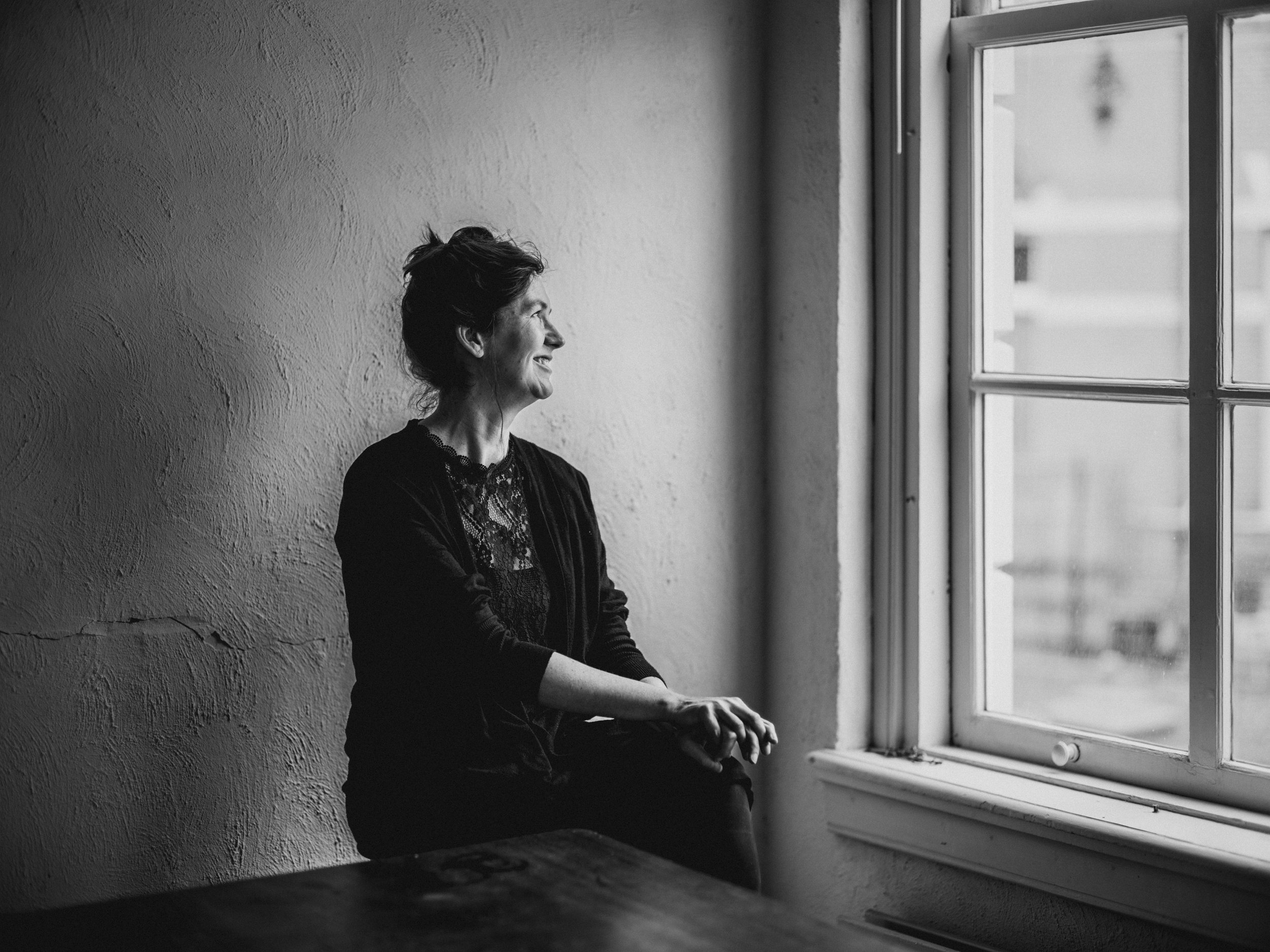 A black and white photo of an older woman with short hair sitting by a window, smiling, looking outward.