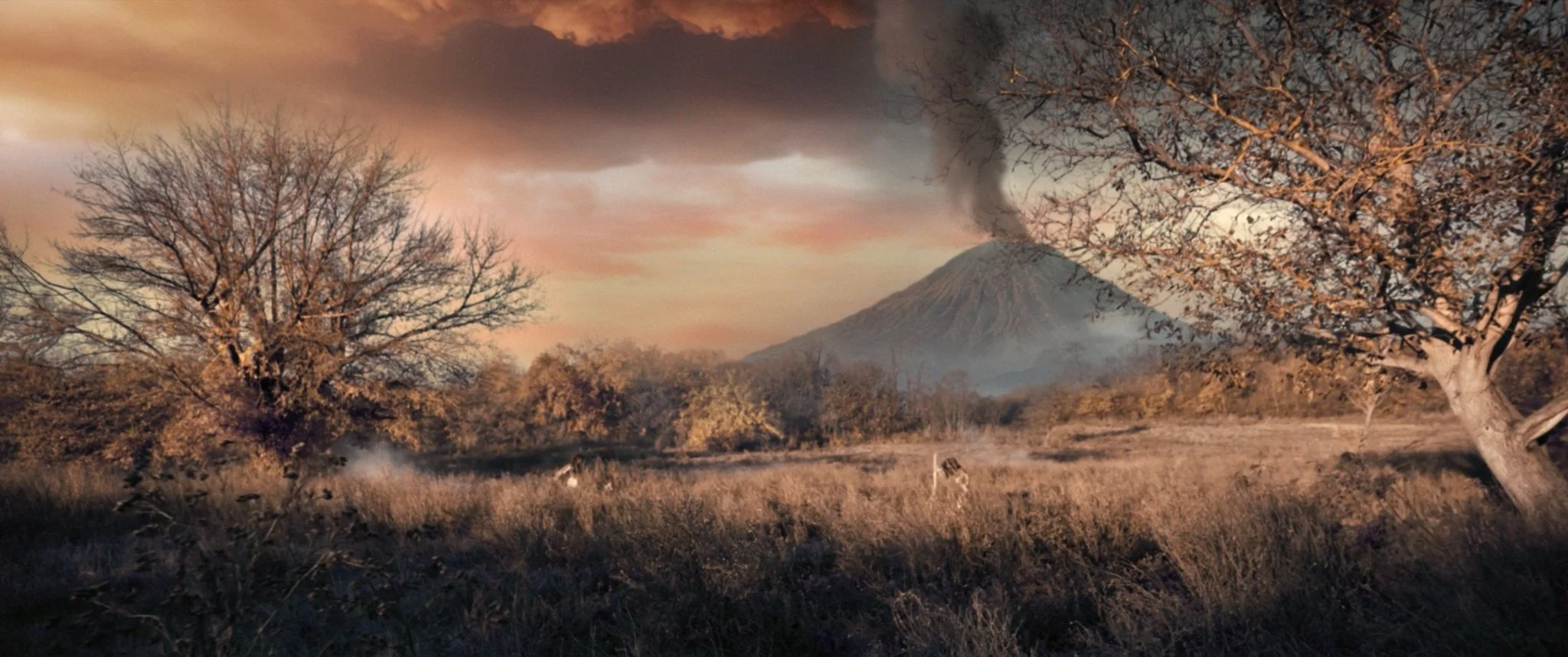 A volcanic eruption with smoke coming from the crater, set against a colorful sunset sky. The scene includes leafless trees and dry grass in the foreground, with distant mountains.
