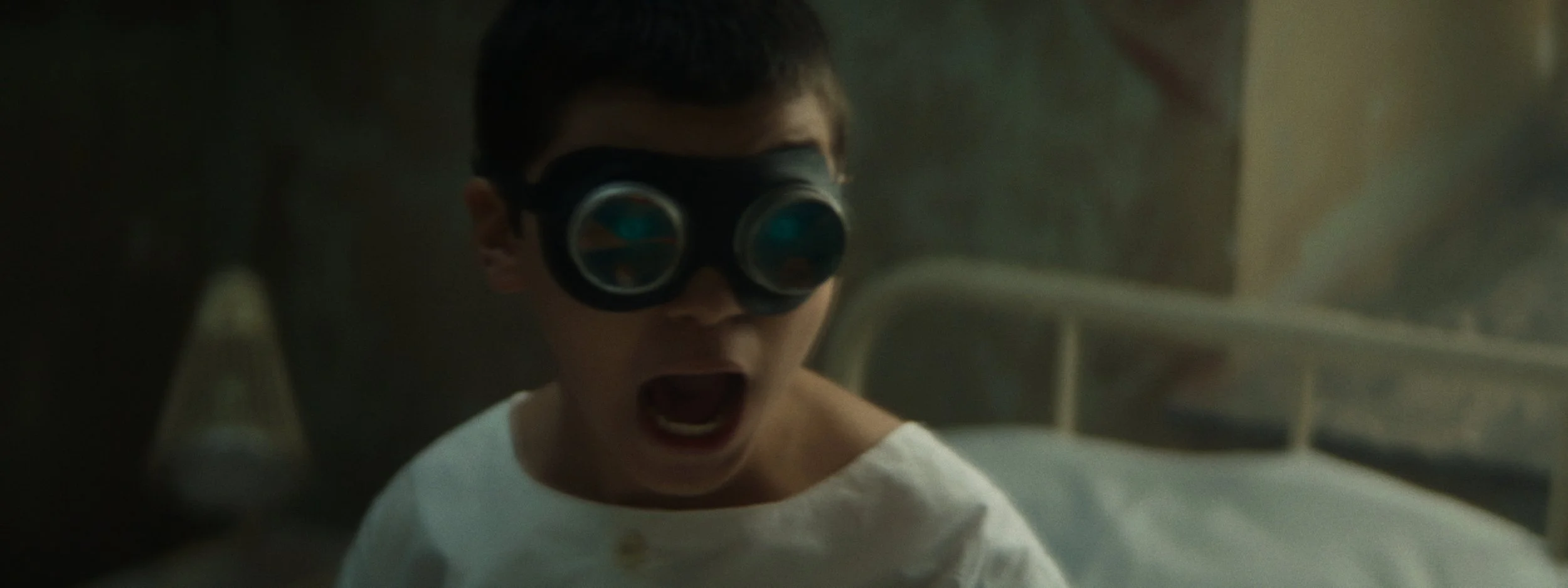 A young boy wearing large, round goggles and a white shirt, with his mouth open in shock or fear, in a dimly lit room with medical equipment in the background.