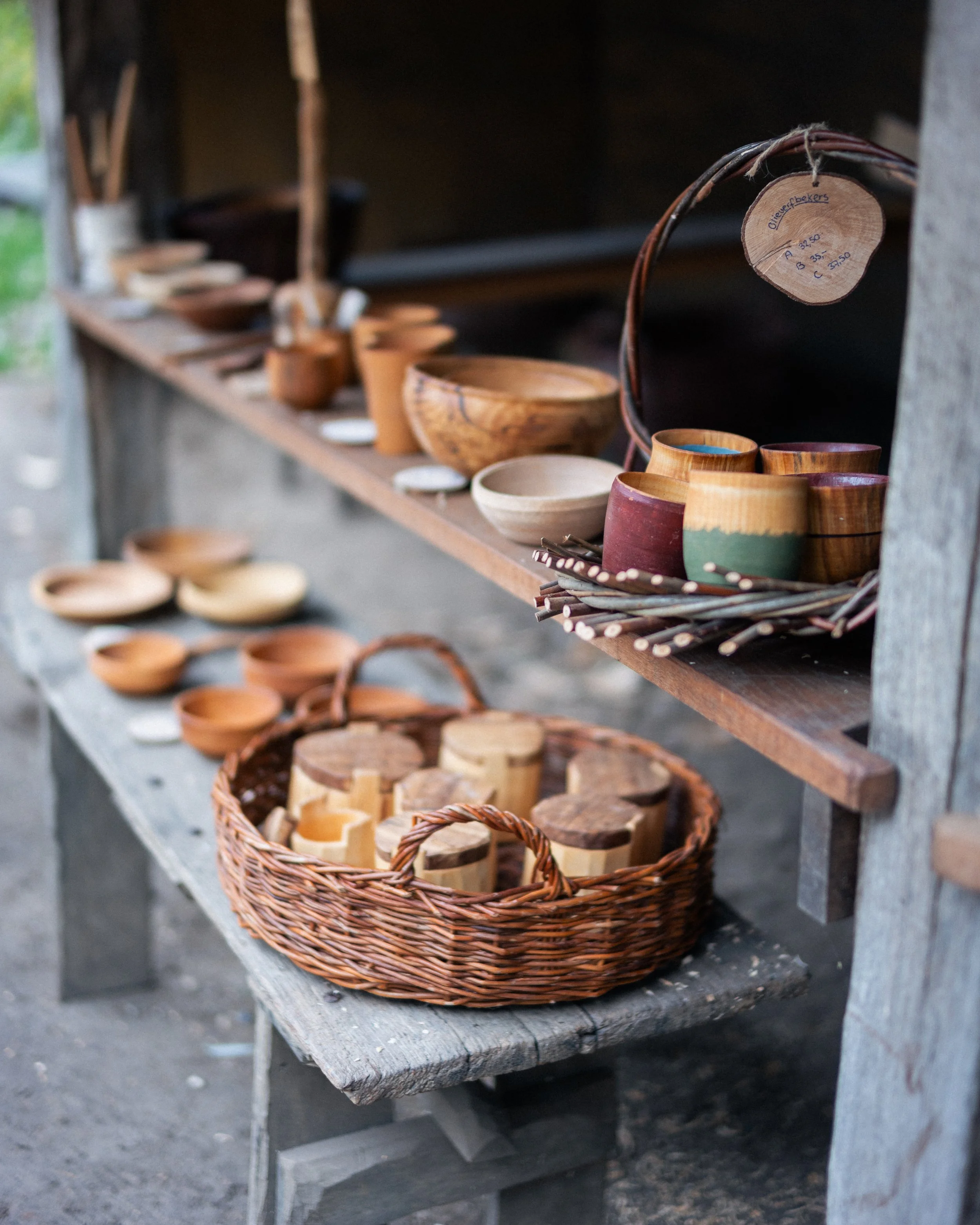 Wooden bowls, cups, and other kitchenware displayed on a rustic wooden shelf and in a woven basket at an outdoor market.