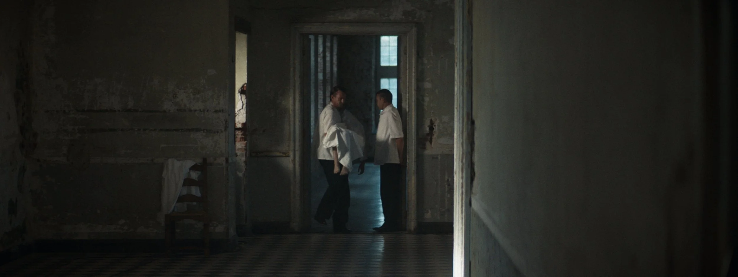 Three people in white coats standing in a dimly lit, rundown interior space, possibly an old building or hospital, with peeling walls and a checkered floor.
