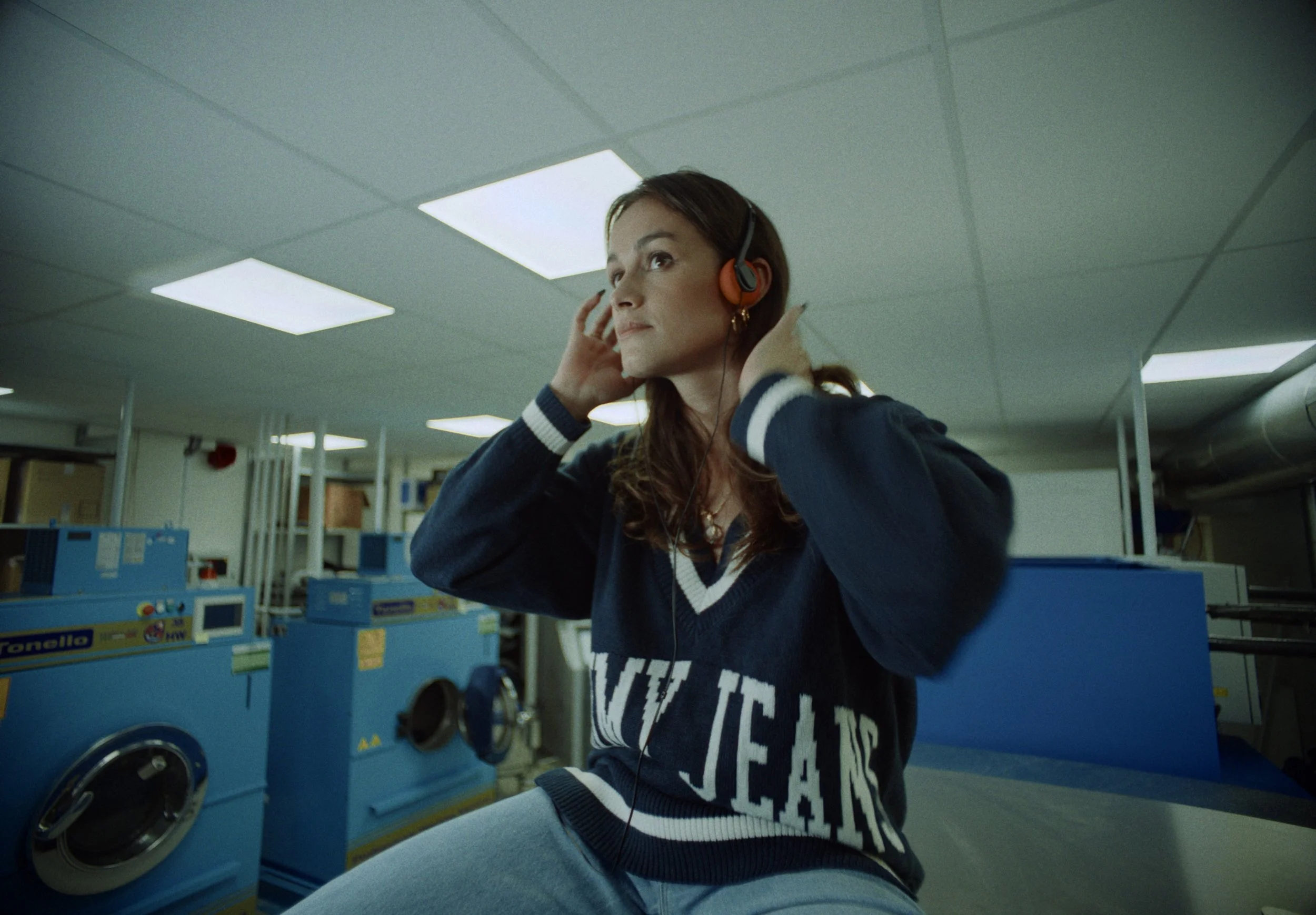 A woman wearing headphones is in a laundromat, adjusting her headphones with her hands near her ears, surrounded by blue washing machines.
