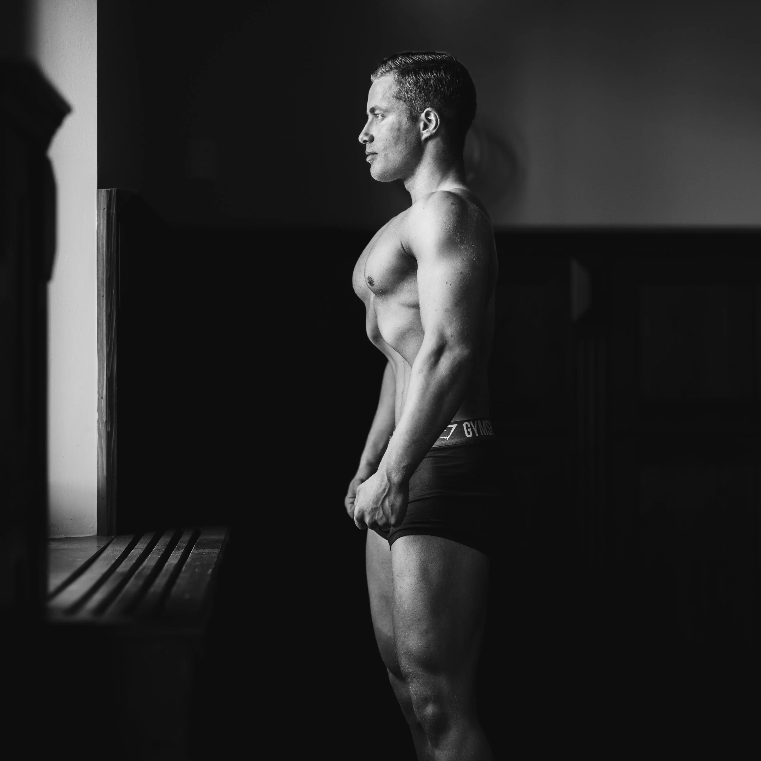 Side view of a shirtless man with short hair standing in a dimly lit room, looking to the right.
