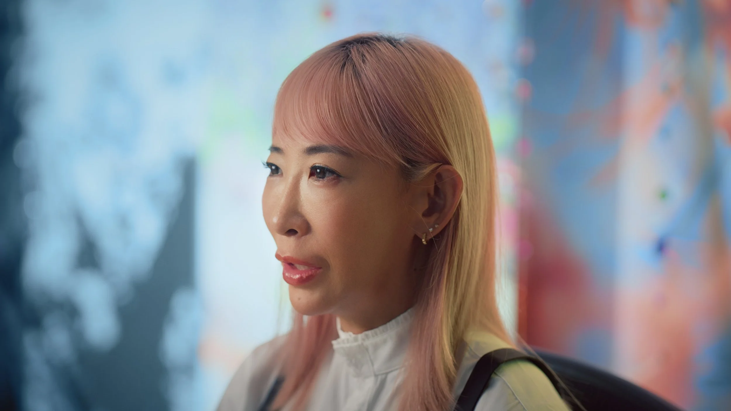 A woman with peach-colored hair and earrings looking to the side, with a blurred colorful background.