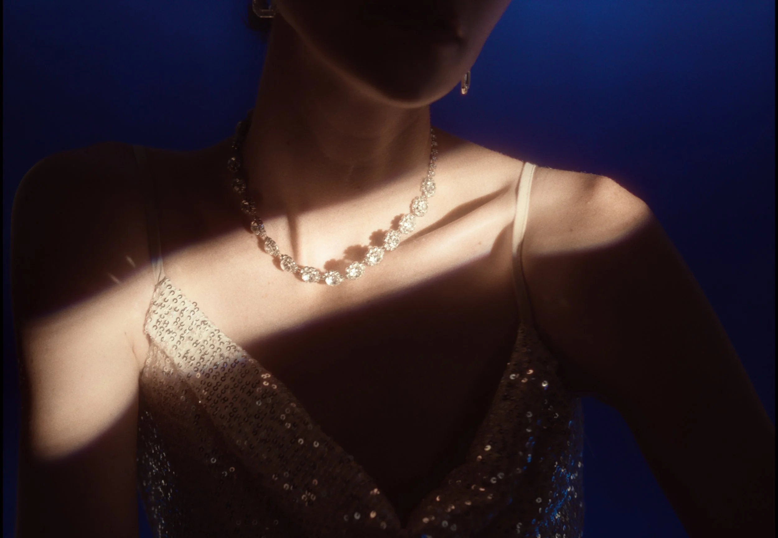 Close-up of a woman wearing a diamond necklace and earrings, with a sequined top, against a dark background.