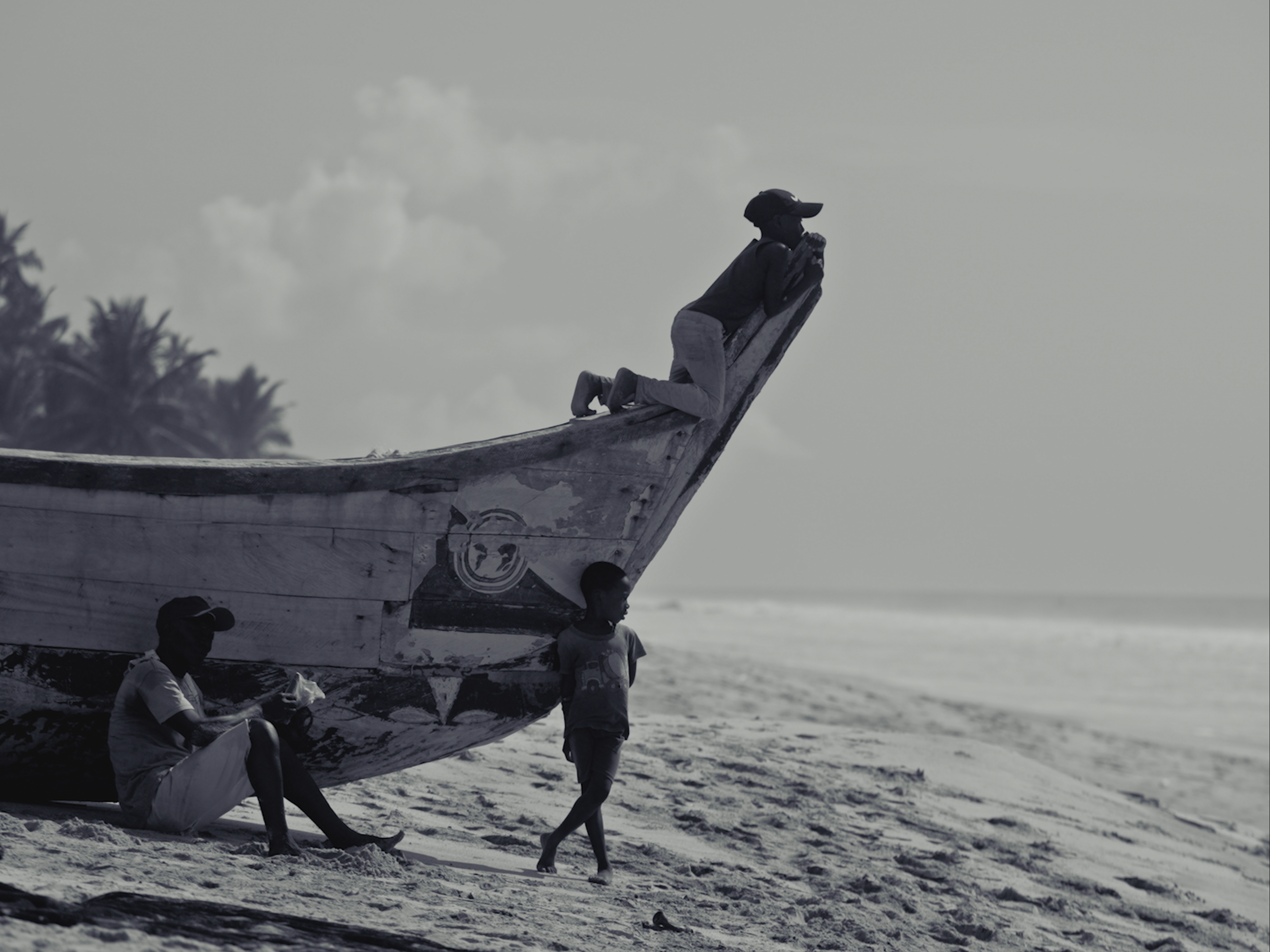 People lifting a boat on the beach, with trees and clouds in the background.
