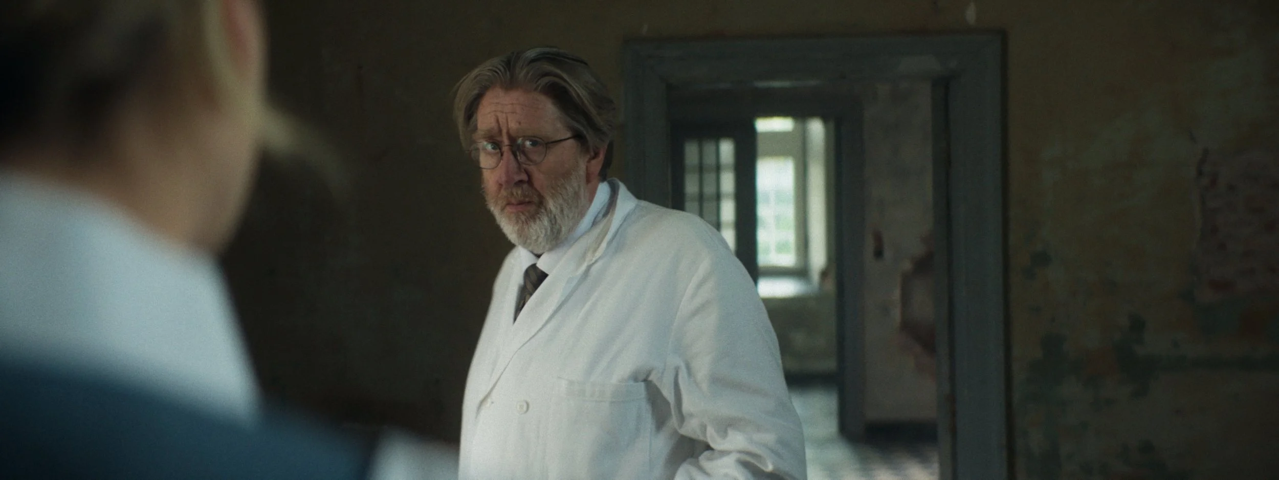A man with glasses and a gray beard wearing a white lab coat, looking surprised, standing in an old, dimly-lit building with exposed brick walls.