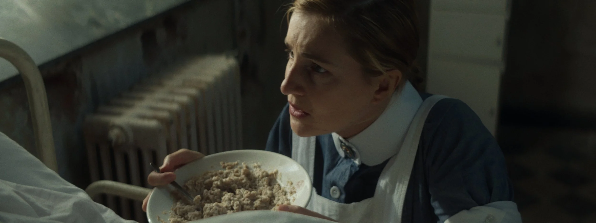 A woman wearing a blue uniform and white apron, sitting on the floor in a kitchen, holding a bowl of mashed food, looking up with an intense expression.