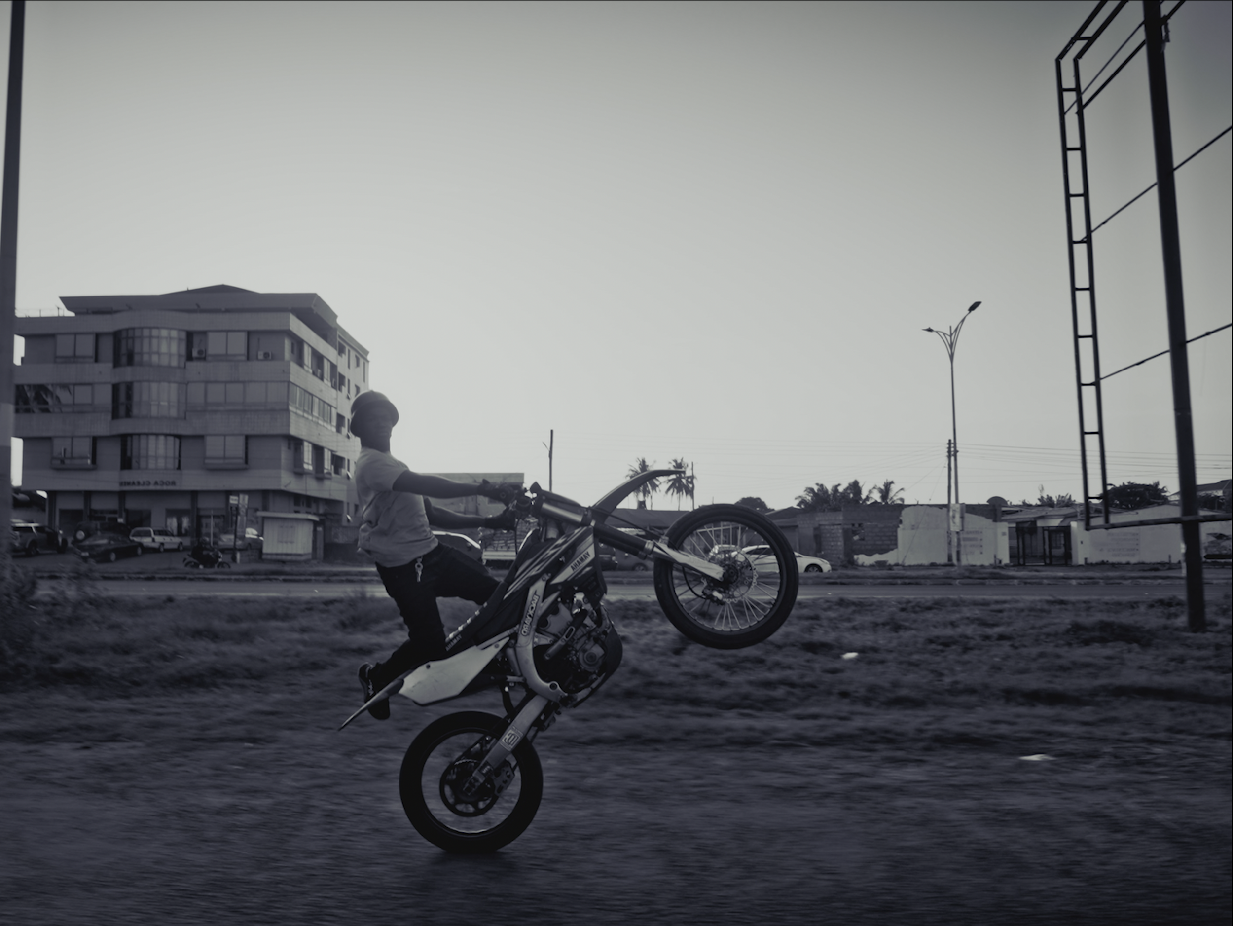 A person performing a wheelie stunt on a dirt bike on a street in an urban area, with buildings, streetlights, and power lines in the background.