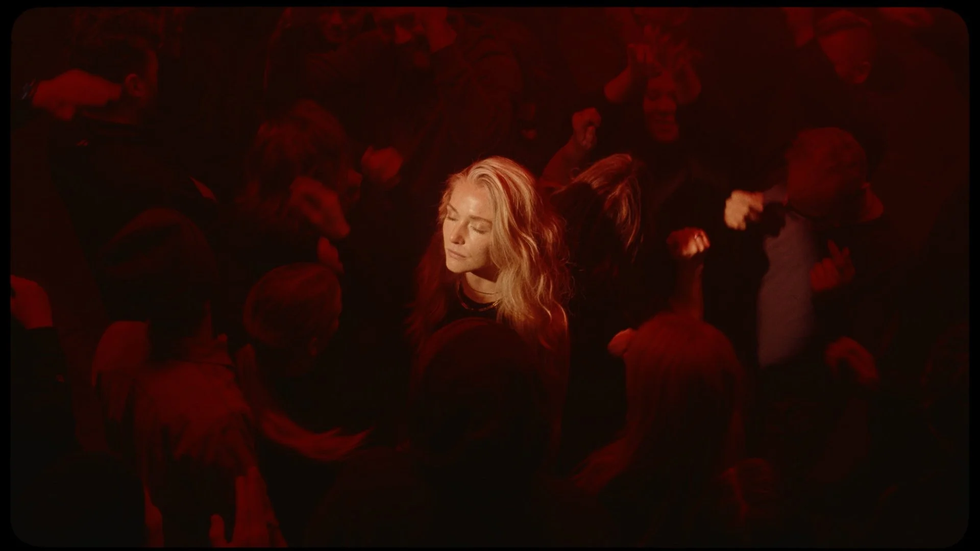 A blonde woman with closed eyes surrounded by a crowd in dim red lighting.