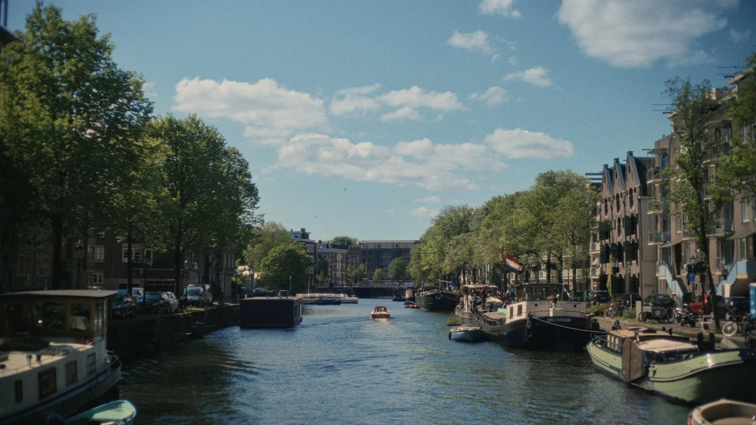 A canal in Amsterdam lined with trees and buildings, with boats docked along the sides and a boat cruising on the water.