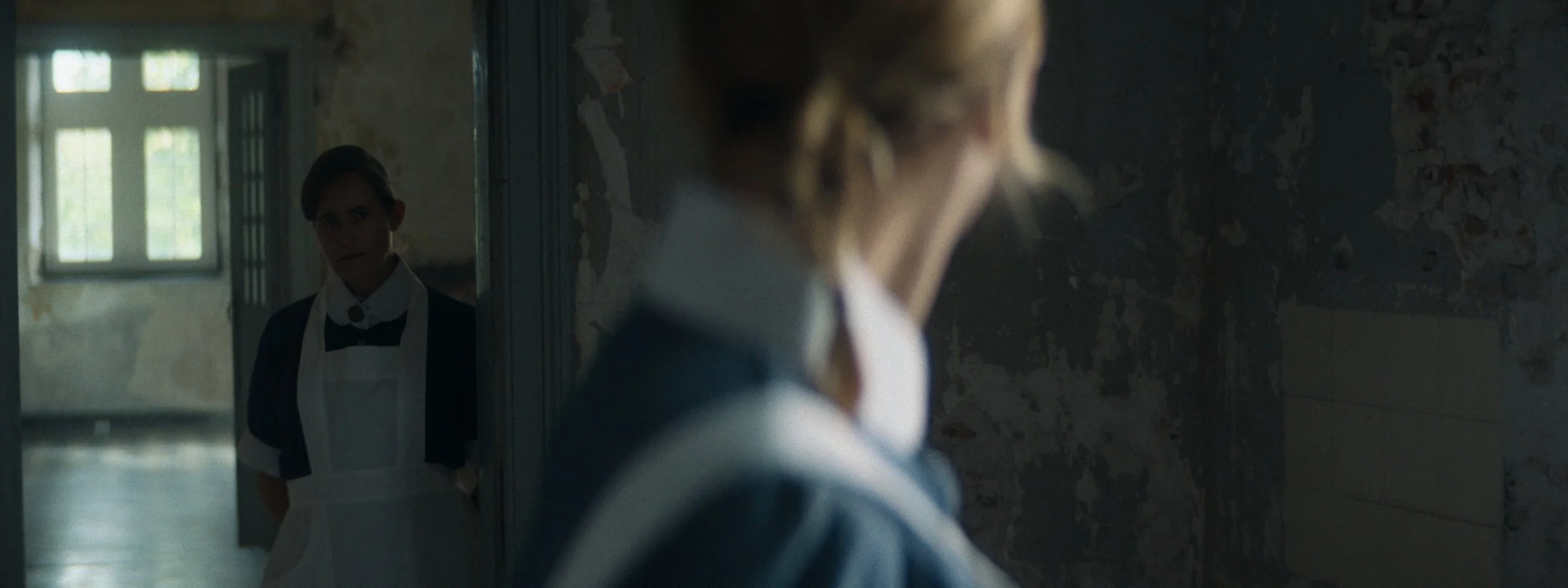 A person with blonde hair viewed in a mirror, with another person wearing a dark uniform and white apron in a dimly lit, worn-down room with peeling walls.