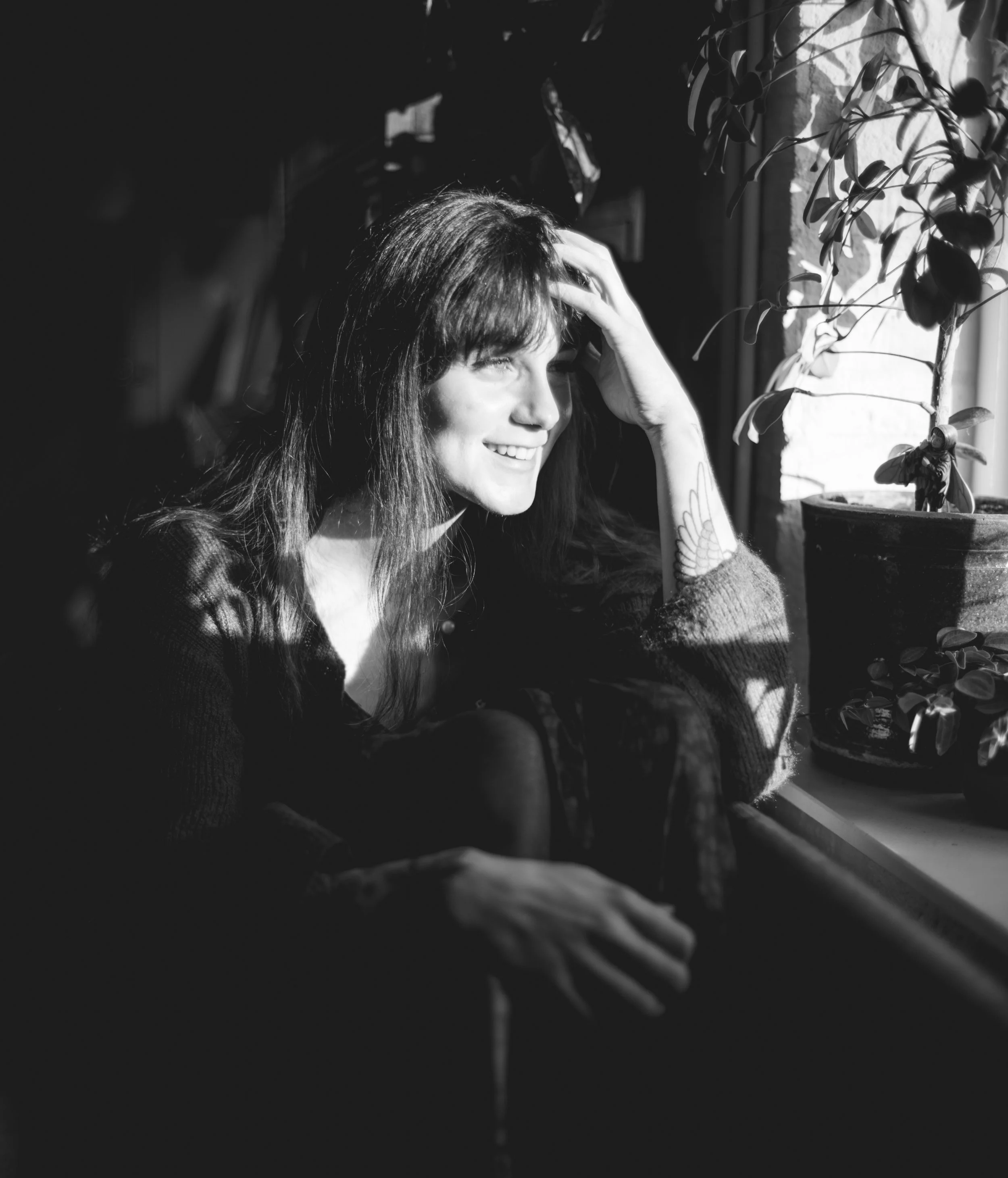 A woman with long dark hair smiling as she looks out a sunlit window, touching her head with her hand. Potted plant with trailing vines is on the windowsill.