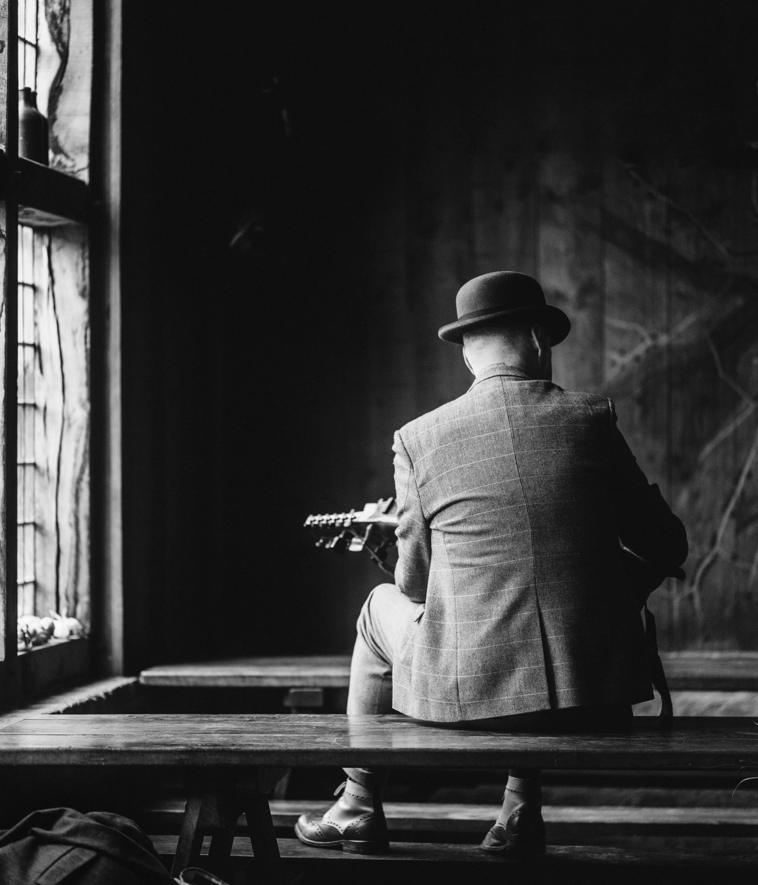 A person wearing a hat and a checkered coat sitting on a bench, playing a guitar, in a dimly lit room with a rustic interior.