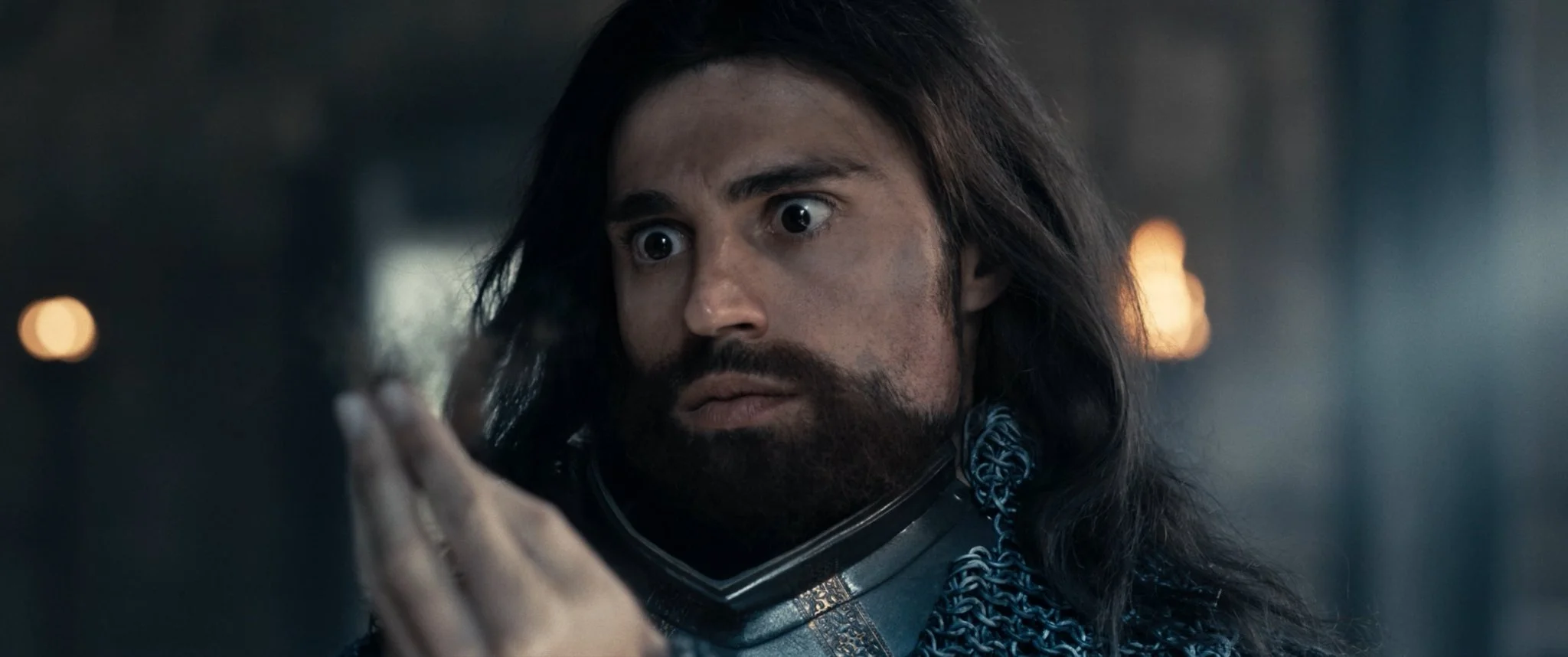A man with long dark hair and a beard, wearing chainmail armor and a space helmet, looks intensely at a device or object in his hand.