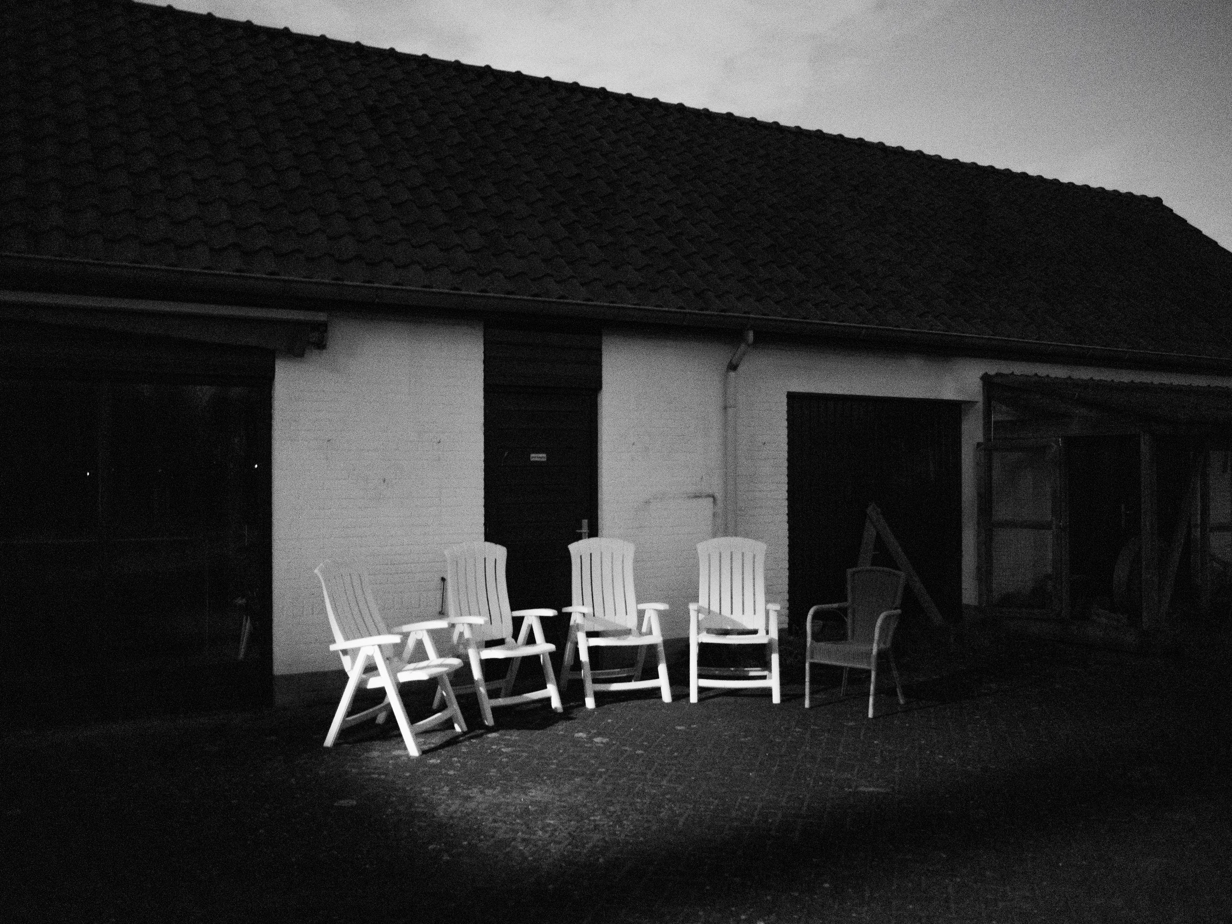 Black-and-white photo of several white plastic chairs arranged outside a house with a tiled roof and a white wall, with some trees or foliage in the background.