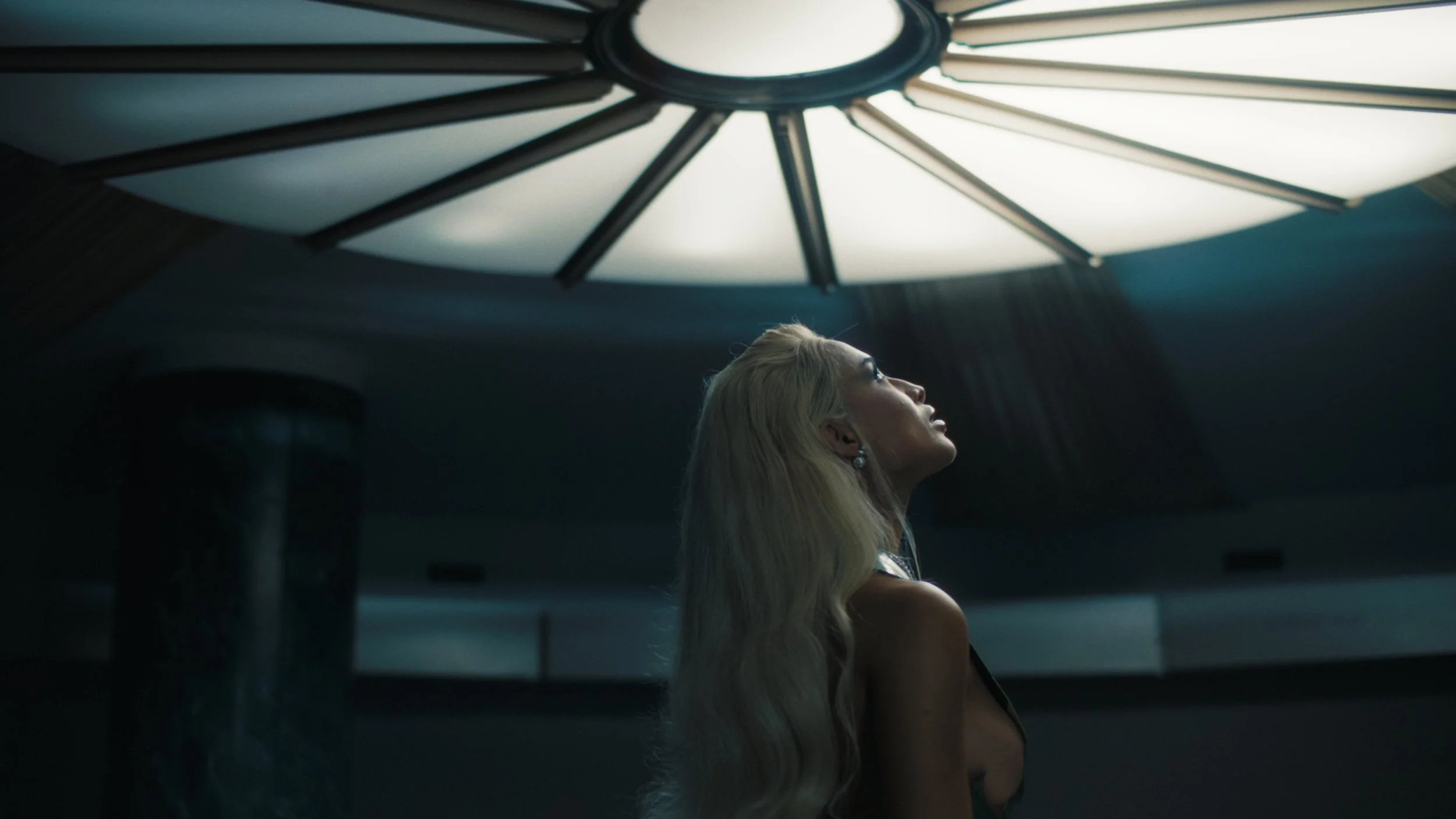 A woman with long blonde hair wearing earrings, looking up at a large, illuminated ceiling light fixture.