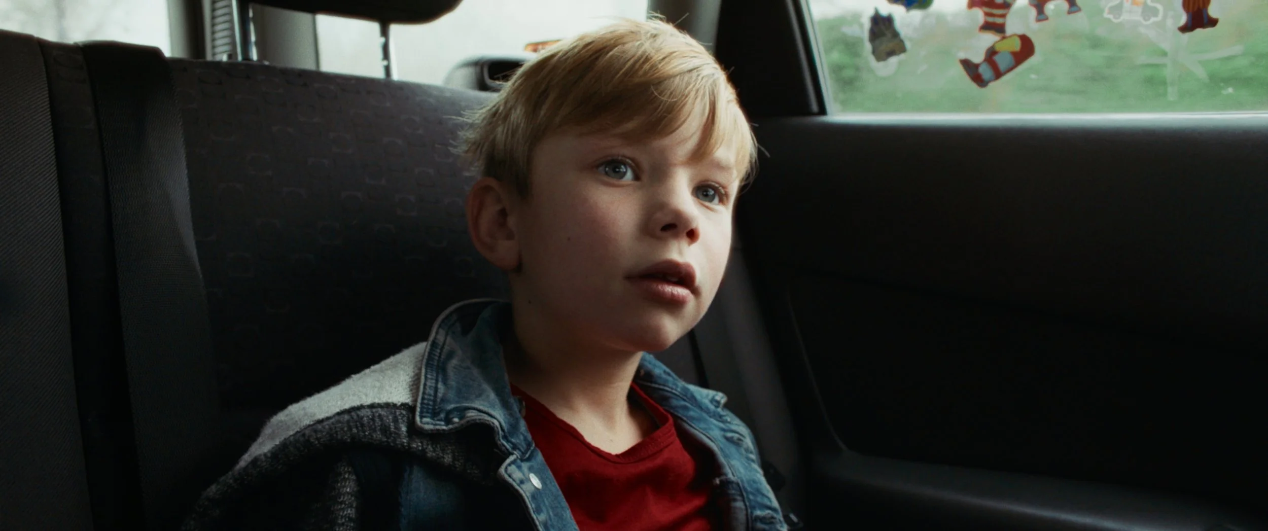 A young boy with blonde hair and blue eyes sitting in the backseat of a car, looking out the window with a thoughtful expression.