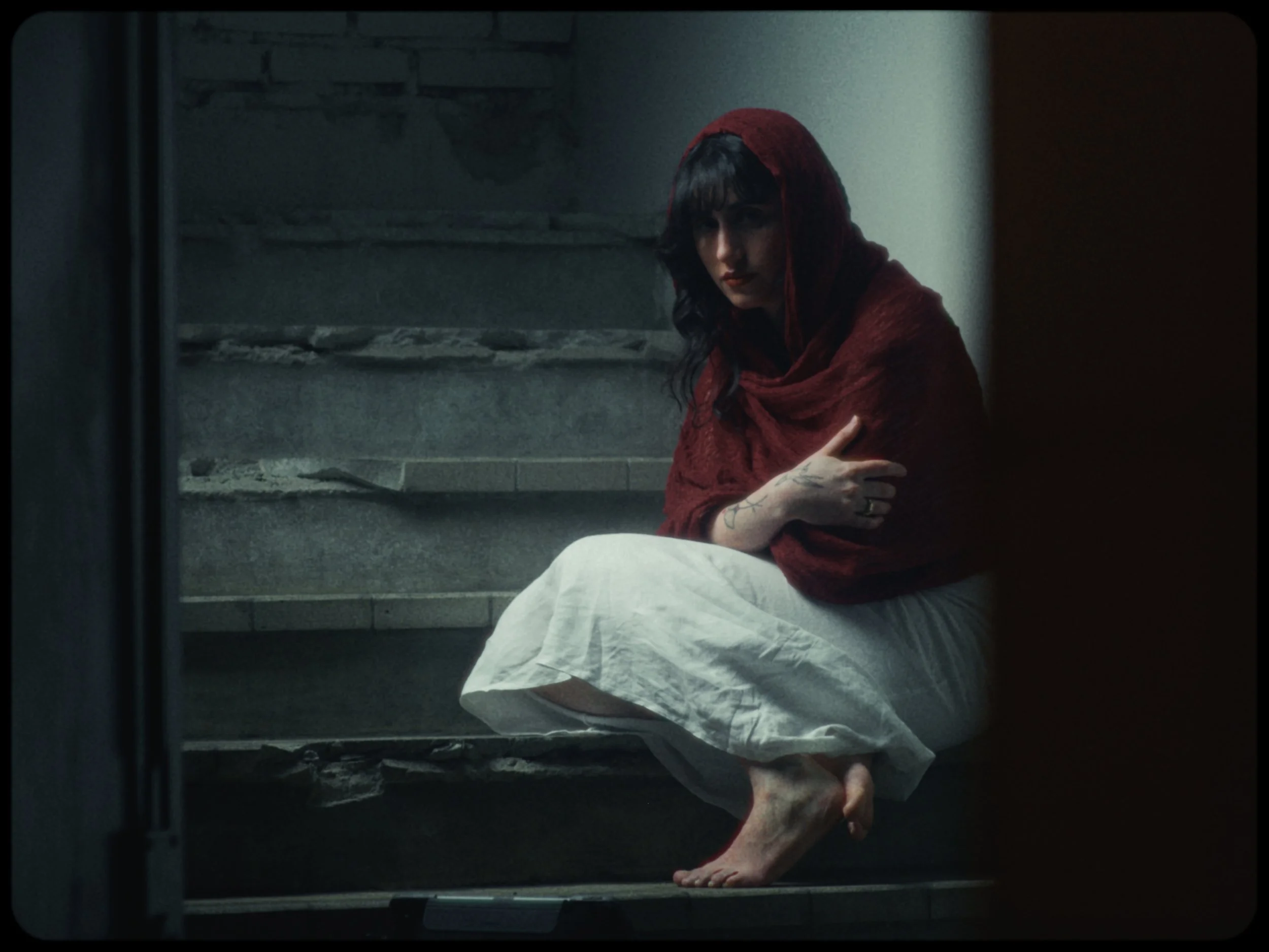 A woman with dark hair and a red hooded garment sitting on the ground in a dimly lit space, with her arms crossed over her knees and tattoos visible on her left arm.