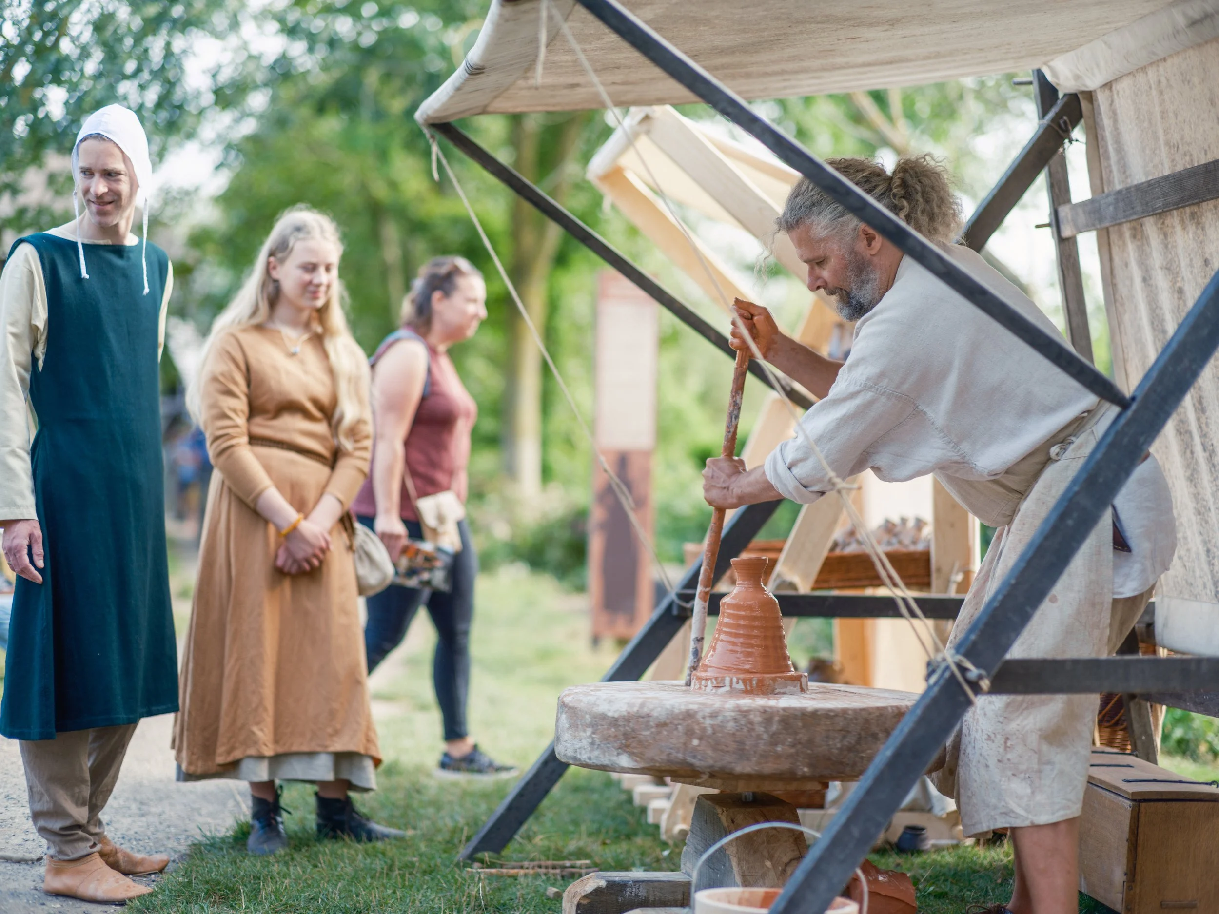 A man and three women at an outdoor pottery demonstration. The man is shaping clay on a potter's wheel, while the women watch. The scene is set in a park with green trees.