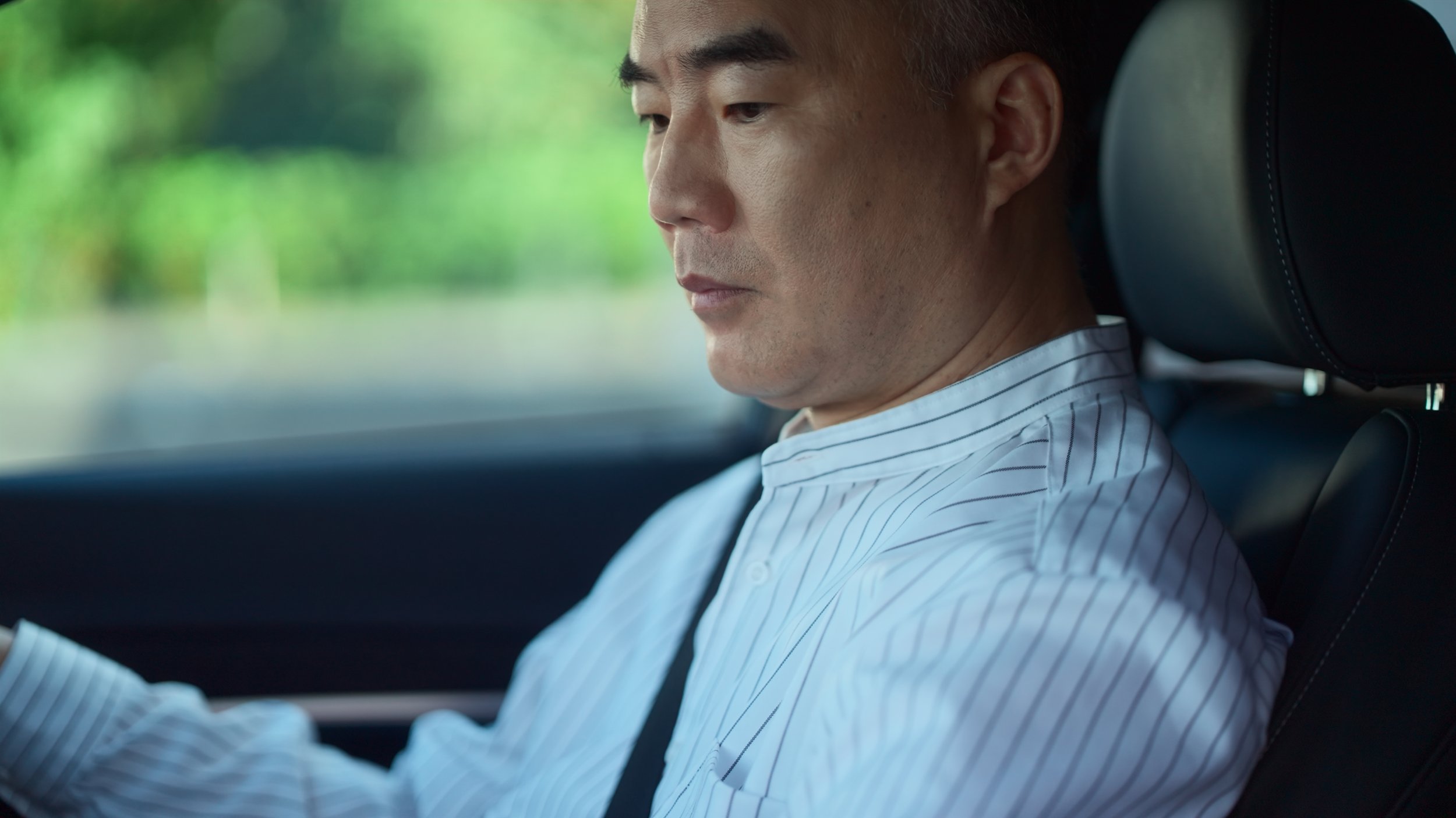 A man with short dark hair and wearing a white striped shirt sitting in the passenger seat of a car, looking down.