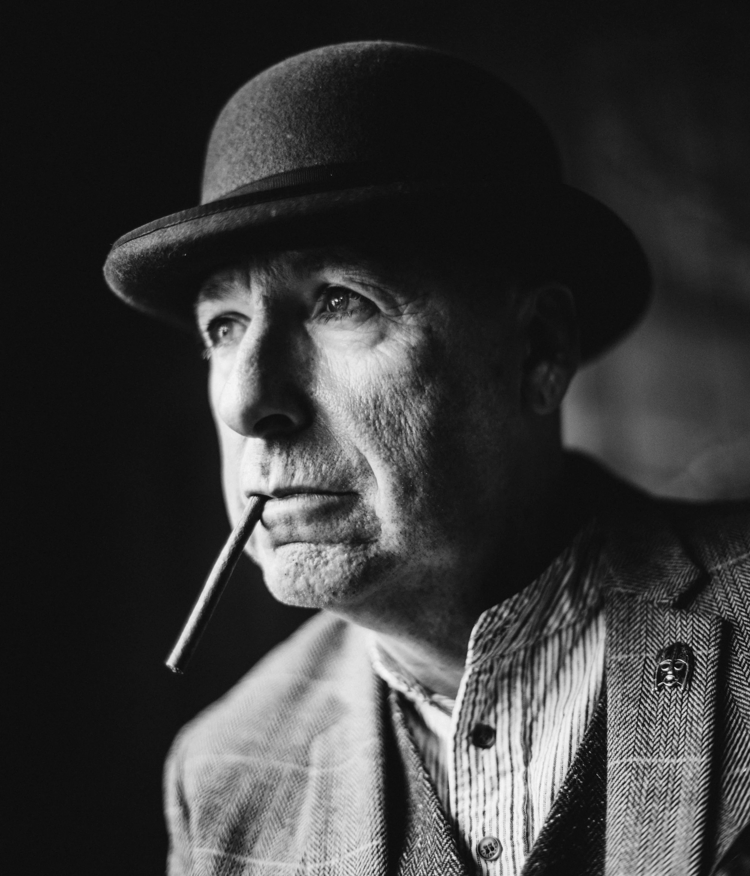 A black and white photo of a man wearing a suit, tie, and patterned dress shirt with a pin. He is wearing a bowler hat and sunglasses, holding a cigarette in his mouth. The image is close-up of his face and upper body.