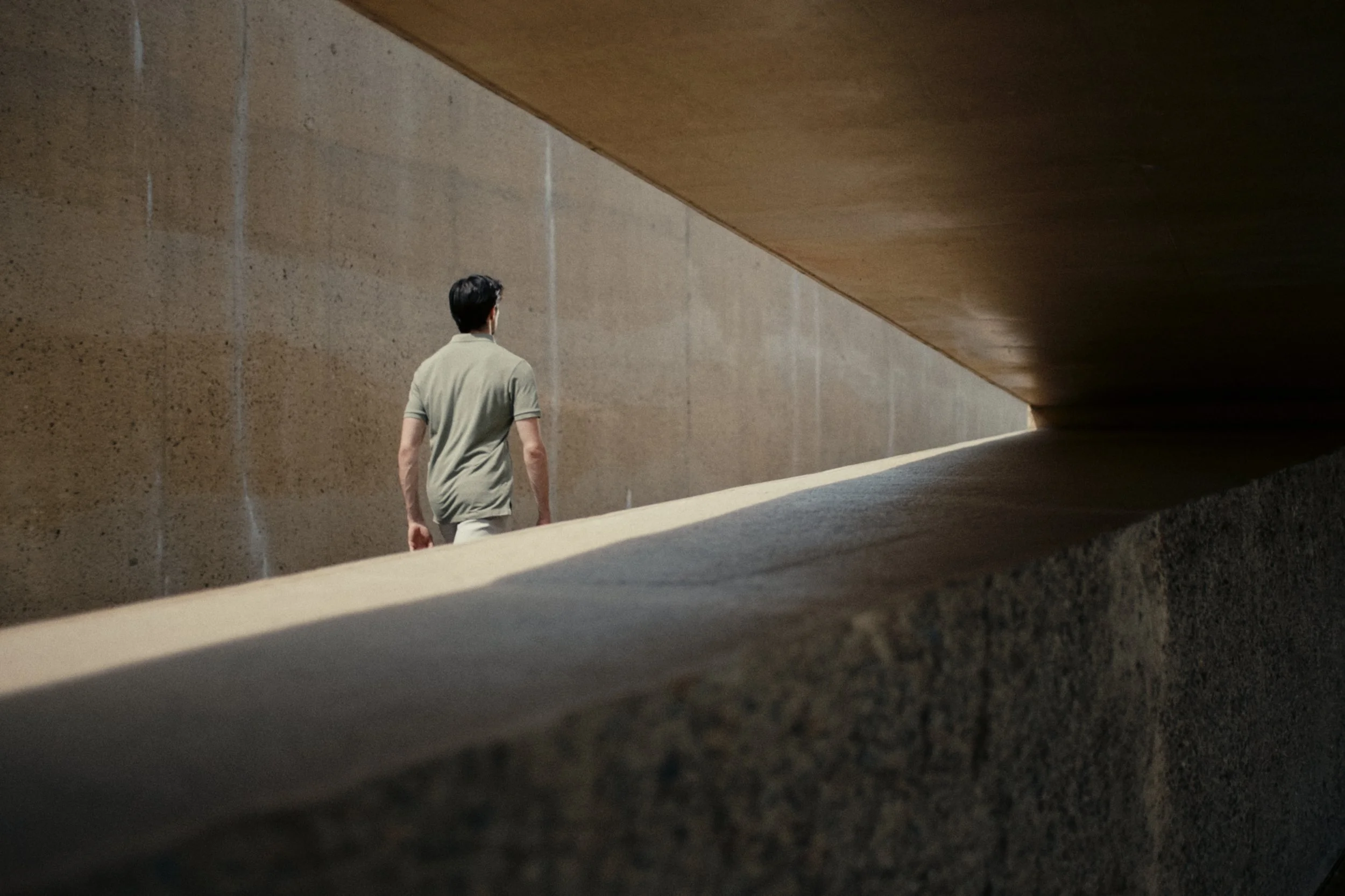 A man with dark hair wearing a beige polo shirt walking along a concrete wall structure, with the underside of a large overhanging concrete surface overhead. The scene appears urban and minimalistic.