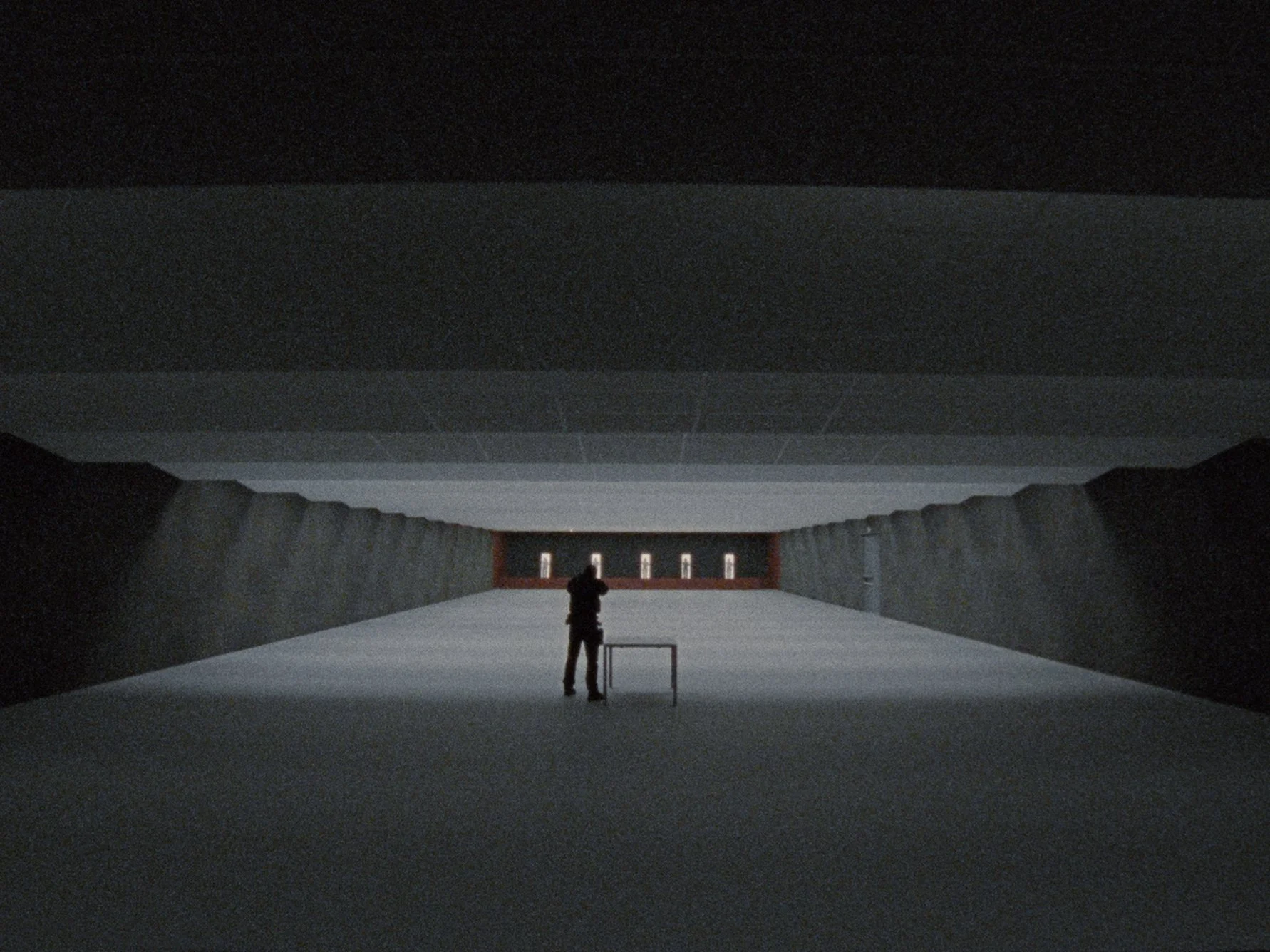 A person standing alone in a spacious, dark, modern interior space with high ceilings and minimal lighting, facing a wall with illuminated windows or display panels.