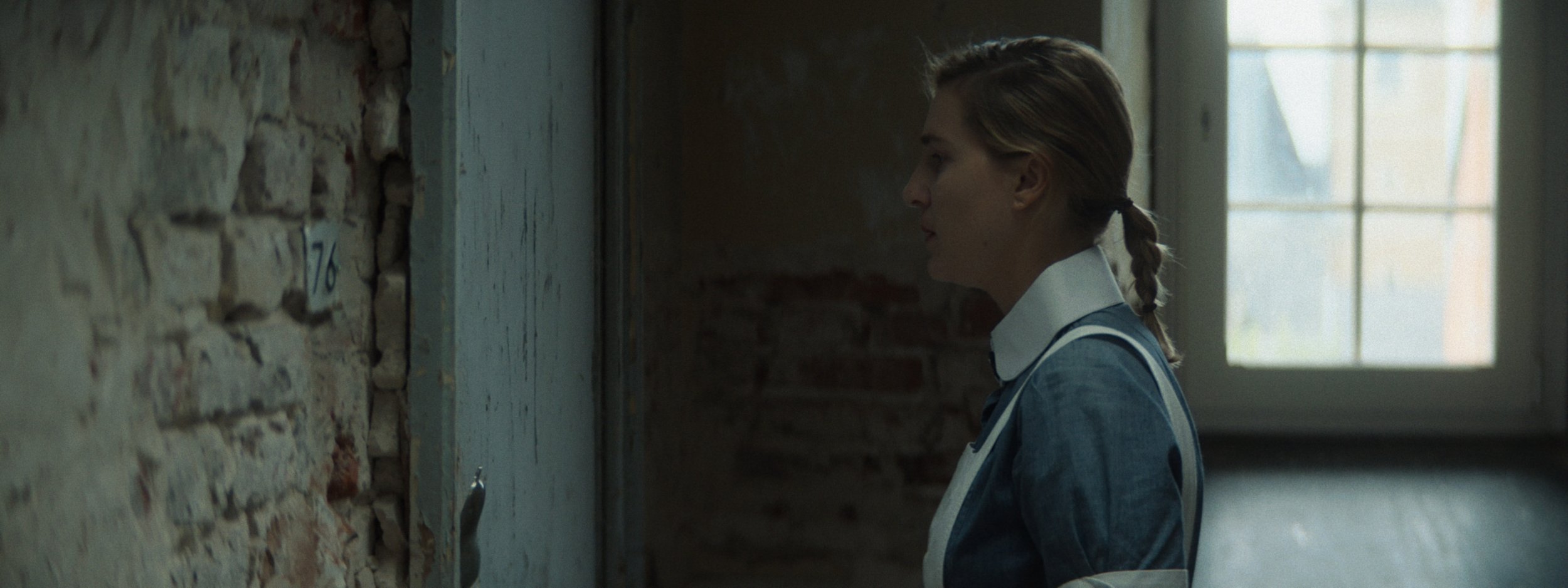 A woman with a braid, wearing a blue uniform with a white collar, standing in profile inside a rustic building with exposed brick walls and a window in the background.