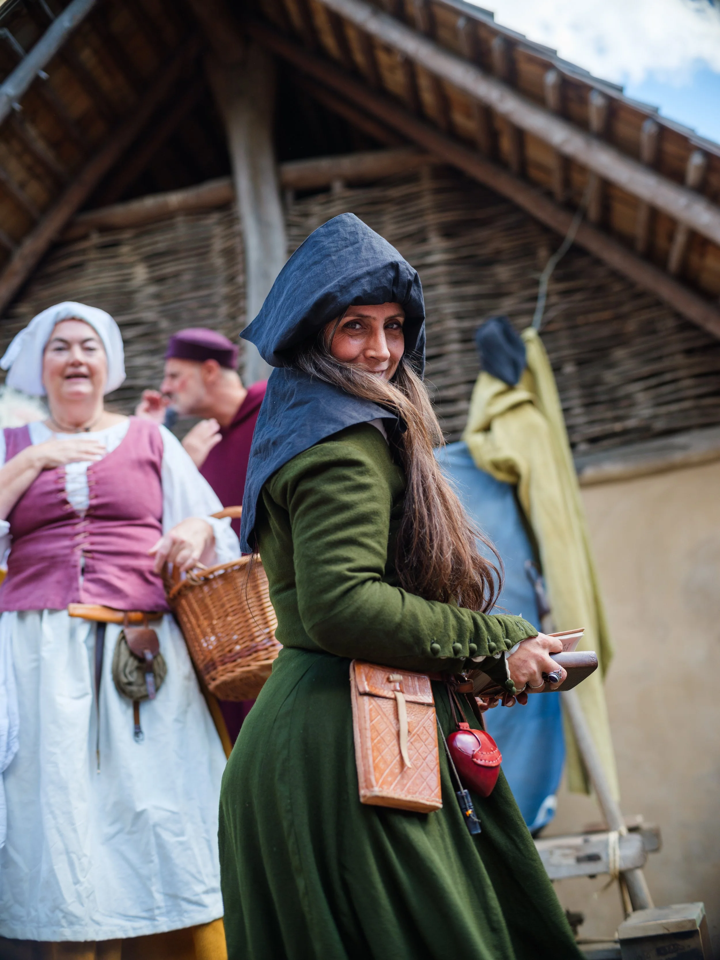 A woman with long brown hair wearing a dark hooded cloak, holding a small book or notebook, standing outdoors under a thatched roof, with other people dressed in historical costumes around her, perhaps participating in a historical reenactment or fes