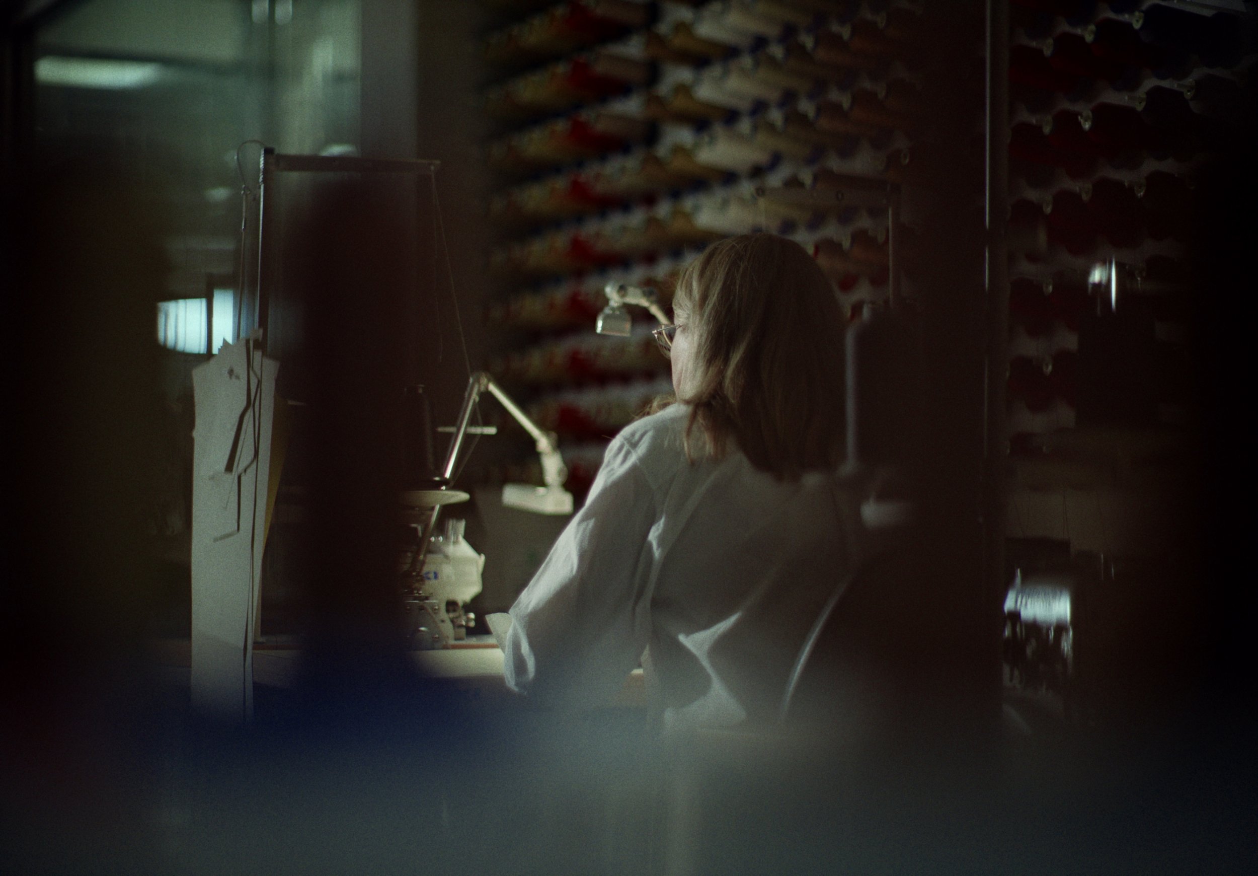 A person with long hair and glasses sitting at a desk in a dimly lit room, possibly a laboratory or workshop, with shelves of supplies or materials behind them.