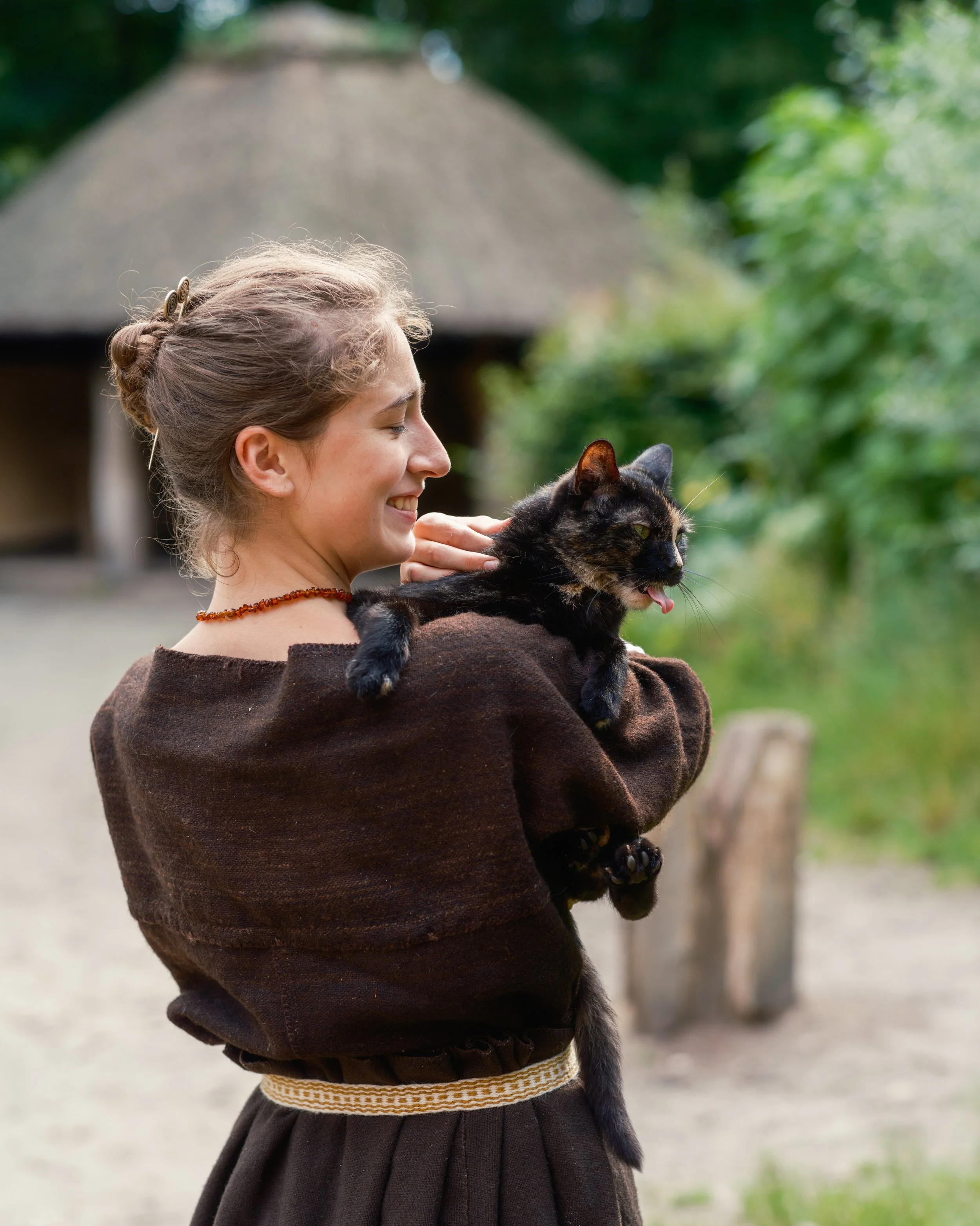 A young girl with light brown hair, styled in a bun with decorative hairpins, is smiling and holding a black and brown cat outdoors. The girl is wearing a dark brown sweater with a striped waistband, and a beaded necklace. The background shows greene