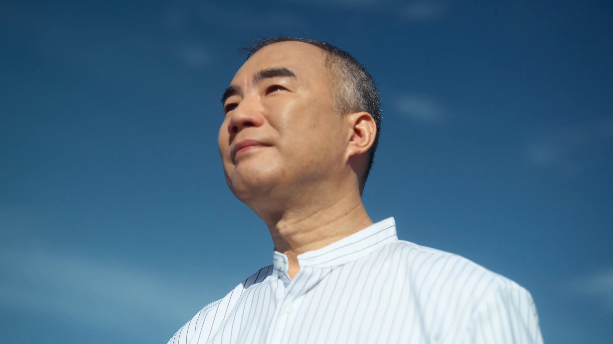 A man with short black hair and a clean-shaven face looking off into the distance, standing outdoors against a clear blue sky, wearing a white shirt with thin vertical stripes.