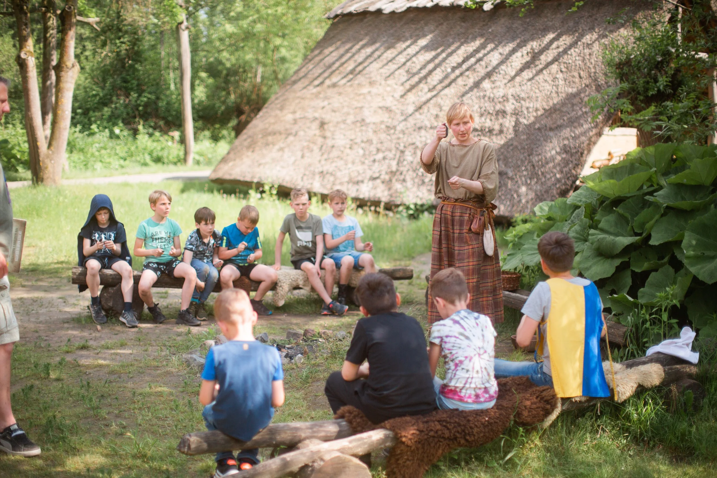 A woman dressed in historical clothing is speaking to a group of children sitting outdoors on benches and logs, surrounded by greenery and a traditional thatched-roof building.