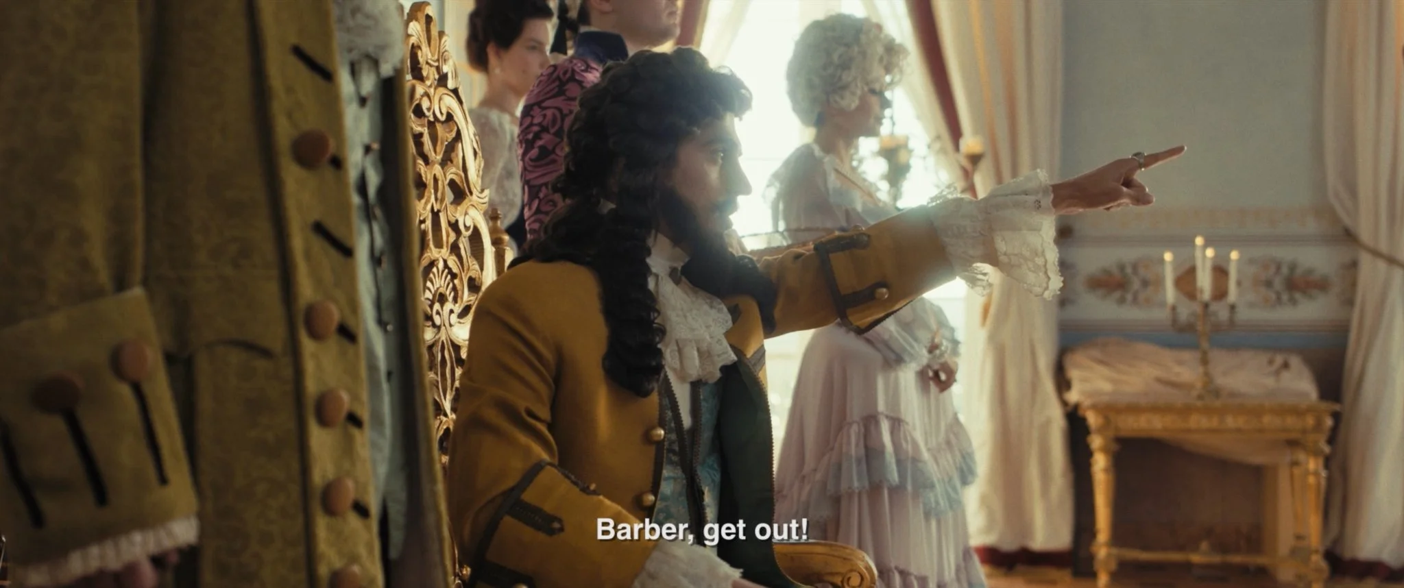 A historical scene with three women and one man dressed in 18th-century clothing in an ornate room. One woman, in a yellow jacket, is sitting on a gilded chair and gesturing with her hand, while another woman in a pastel dress stands nearby. The scen