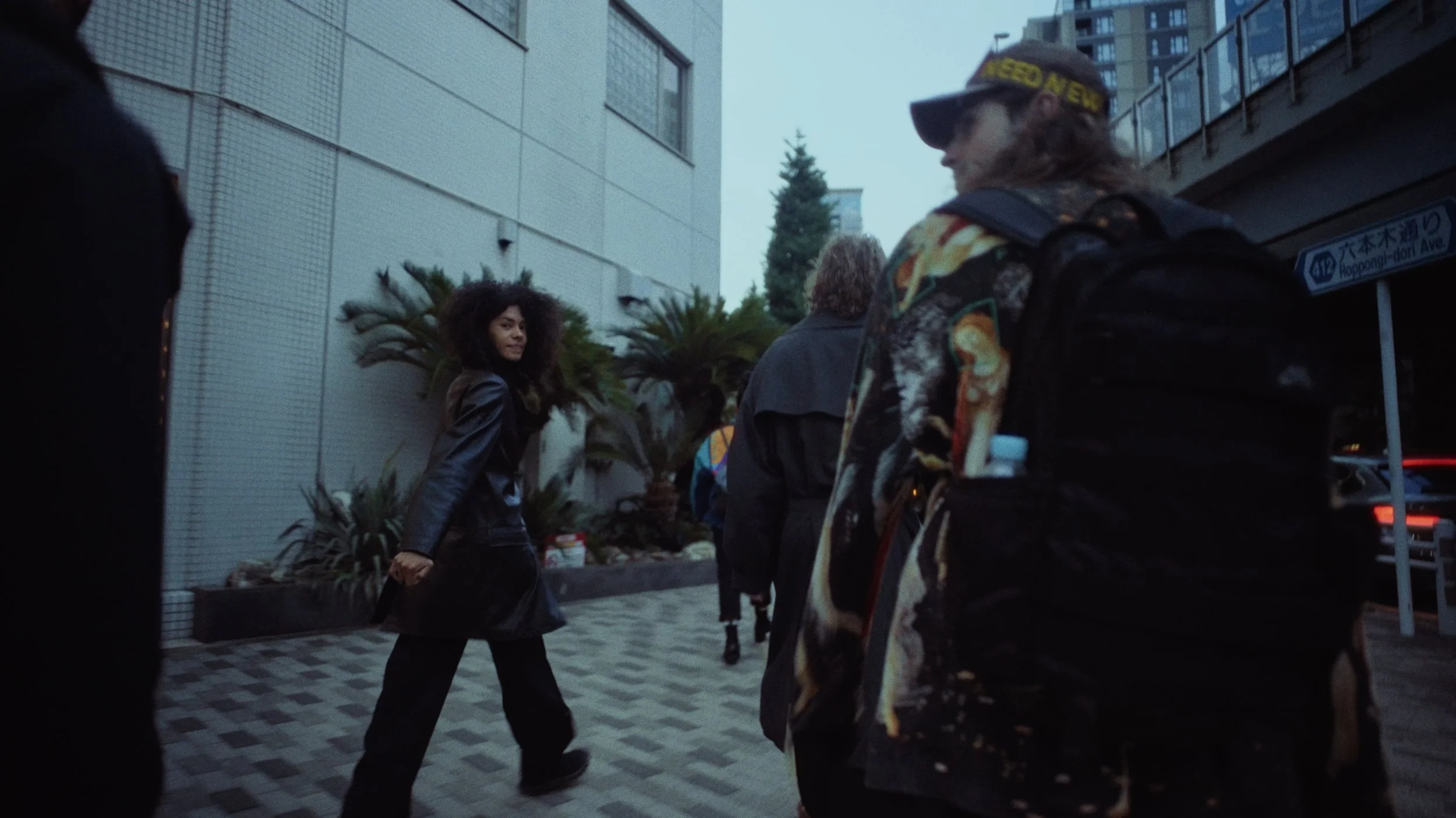 A woman with curly hair wearing a black leather jacket walking on a city sidewalk and looking towards her left, while a group of people, including a woman with a backpack, walk ahead of her. The background shows tall buildings and trees.