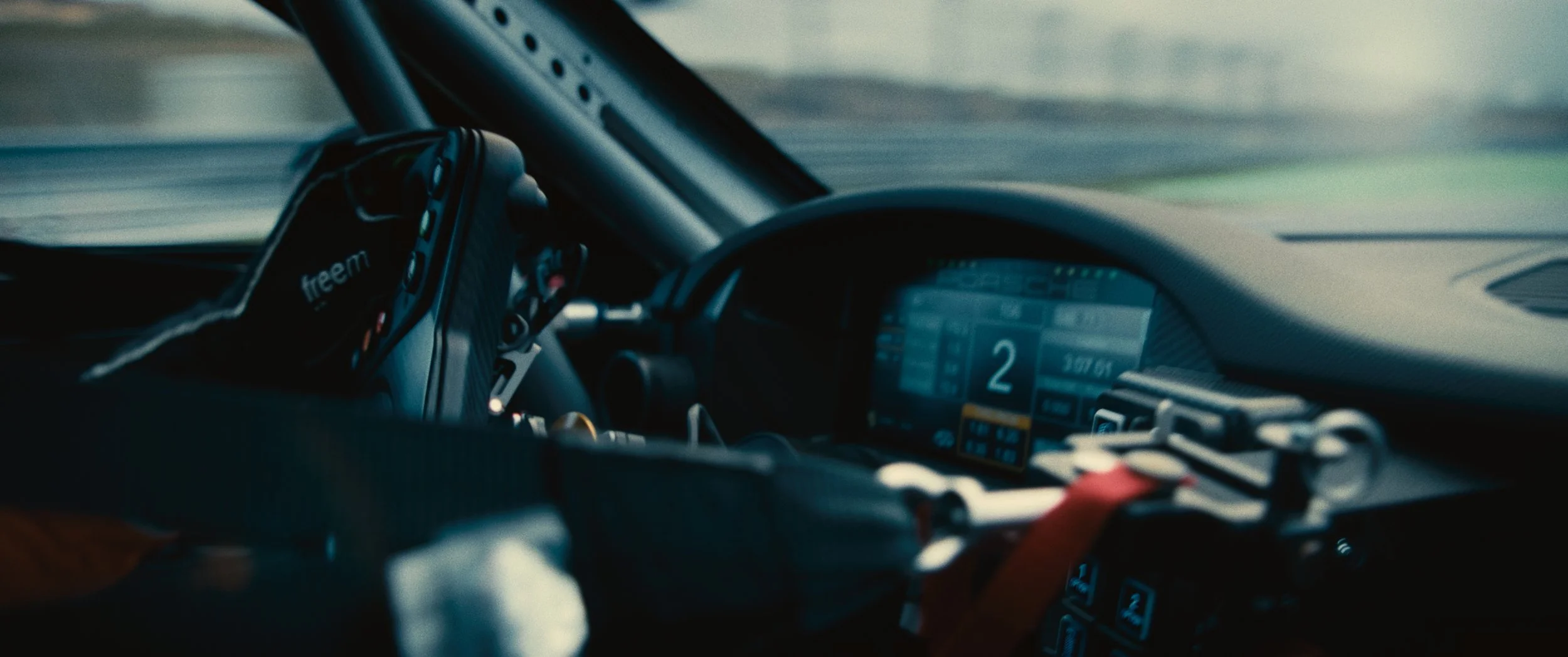Interior of a race car showing a digital dashboard display, steering wheel, and multiple electronic devices, with a blurred view of the racetrack outside.