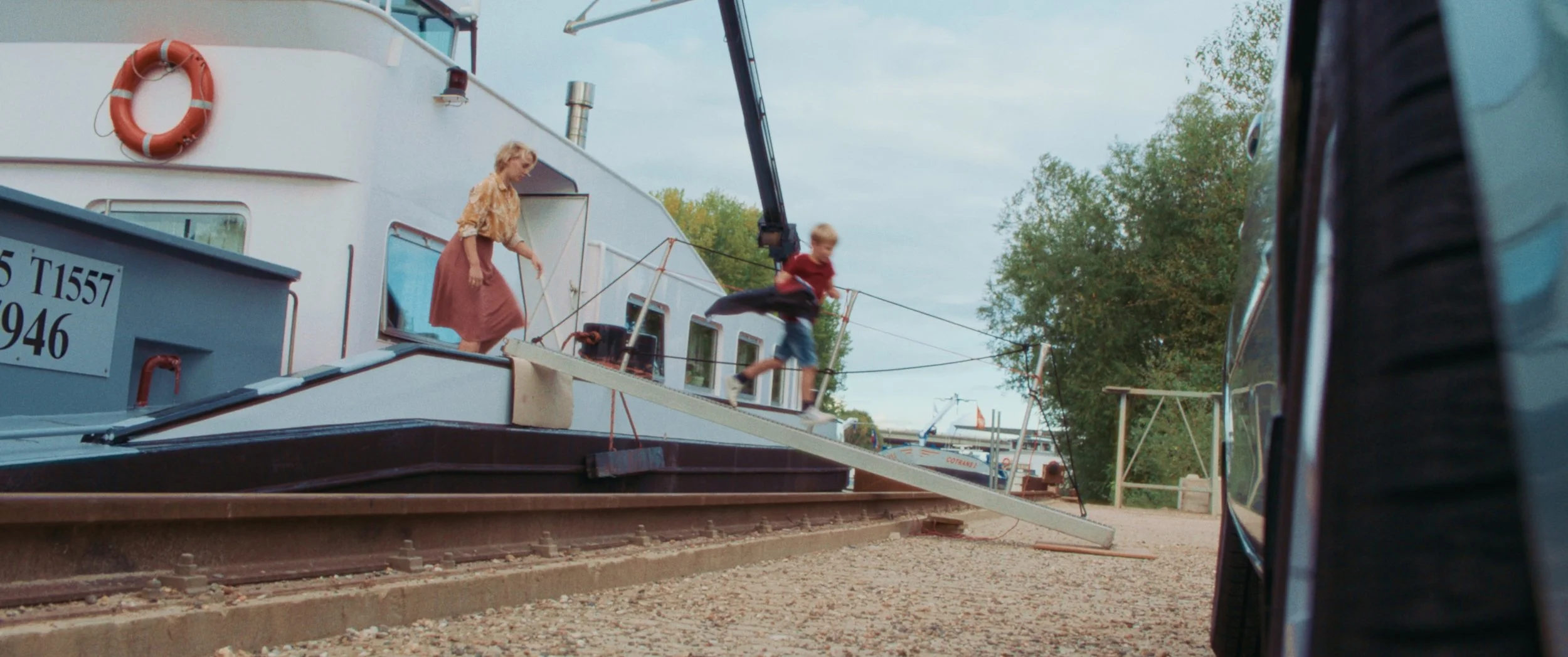 A woman and a boy are on a boat with a gangplank extending to the land. The boy is running across the gangplank while the woman stands on the boat. There are trees and other boats visible in the background.