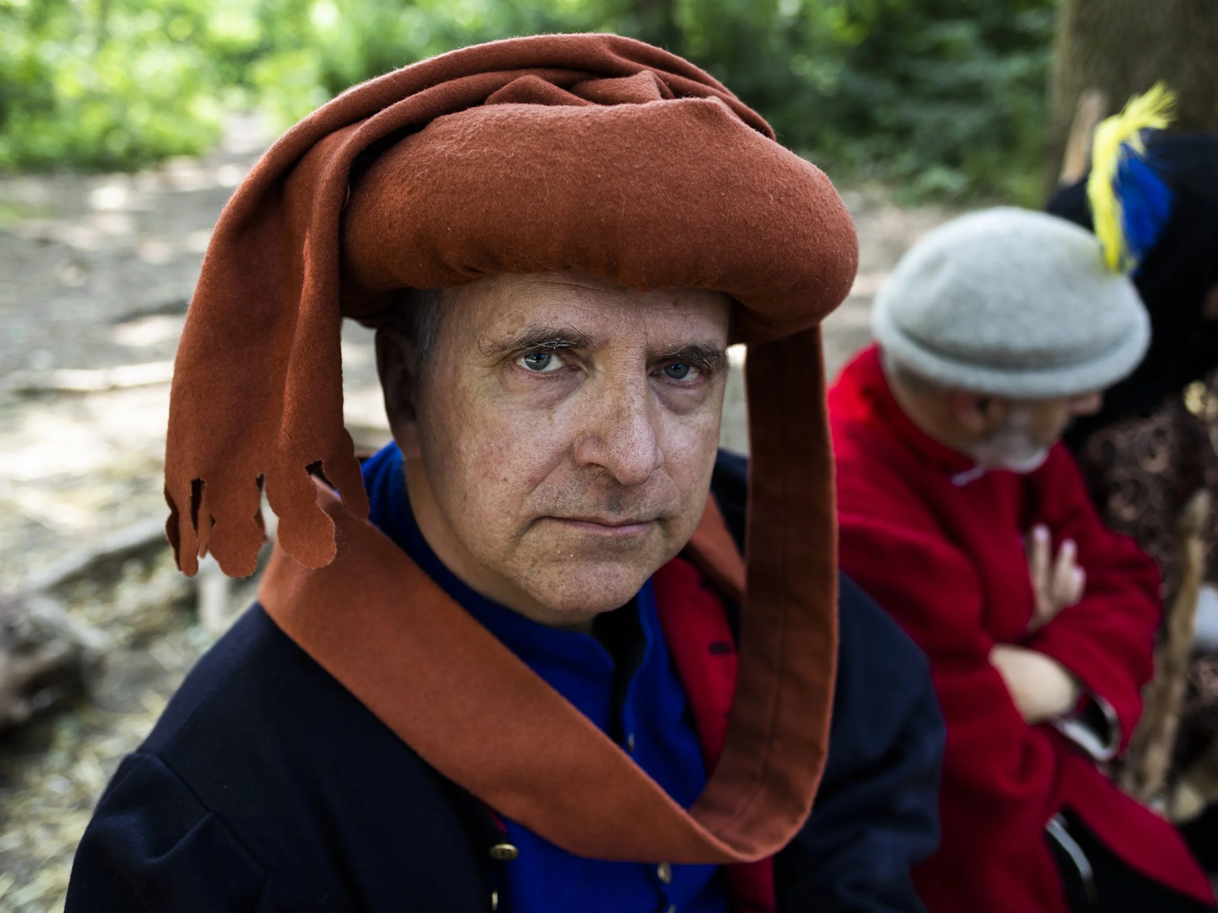 A man wearing a large reddish-brown hat with drooping sides, sitting outdoors with other people in the background, surrounded by trees.
