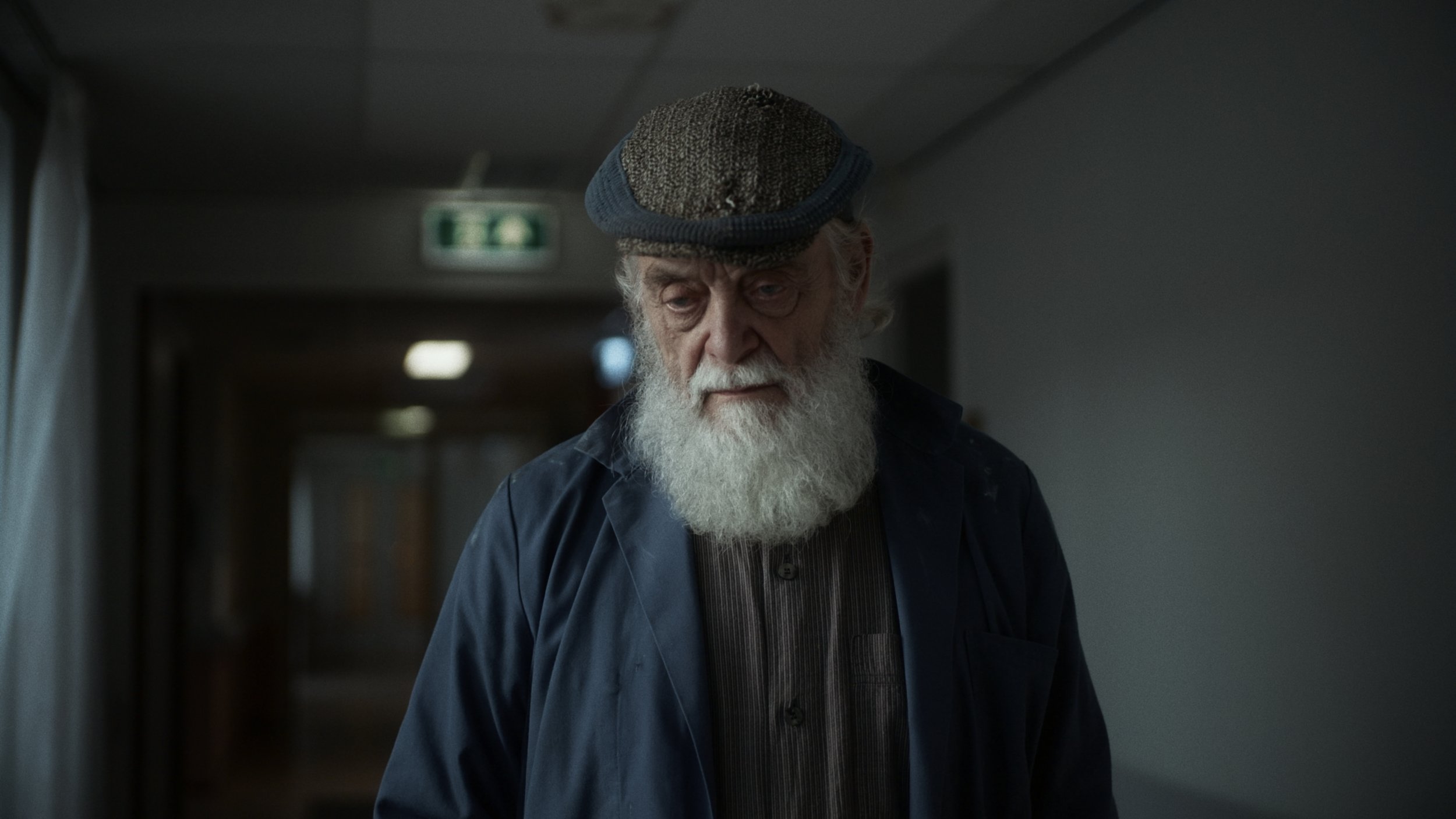 An elderly man with a white beard and glasses walking down a dimly lit hallway, wearing a blue cap and dark clothing.