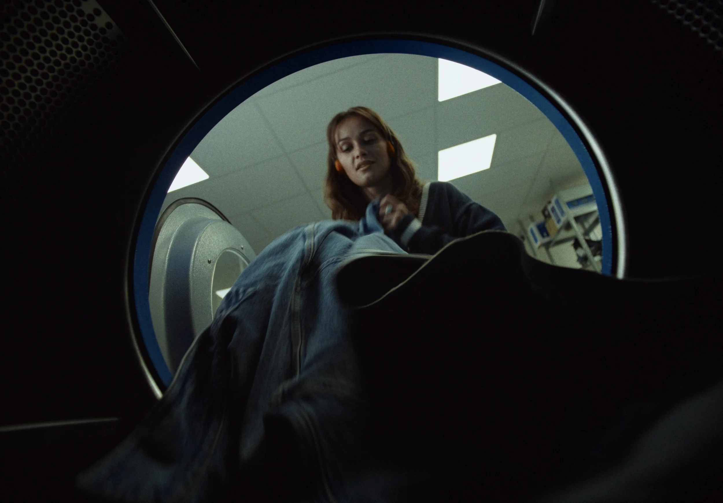 A woman wearing orange headphones is seen through the opening of a washing machine or dryer, looking down at clothes inside.