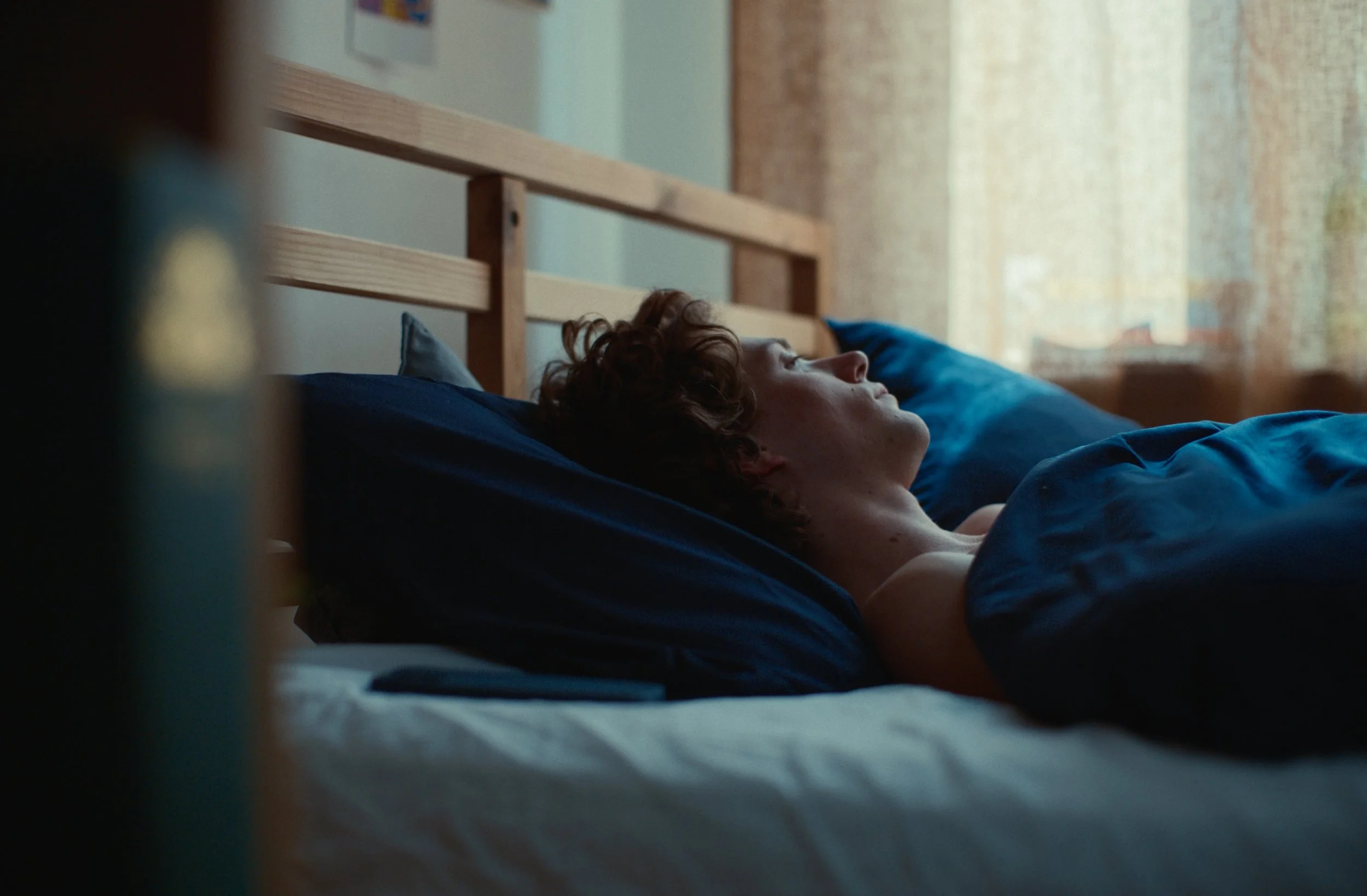A person with curly hair sleeping on a bed with blue sheets, viewed from a low angle, with a wooden headboard and sunlight coming through a window in the background.